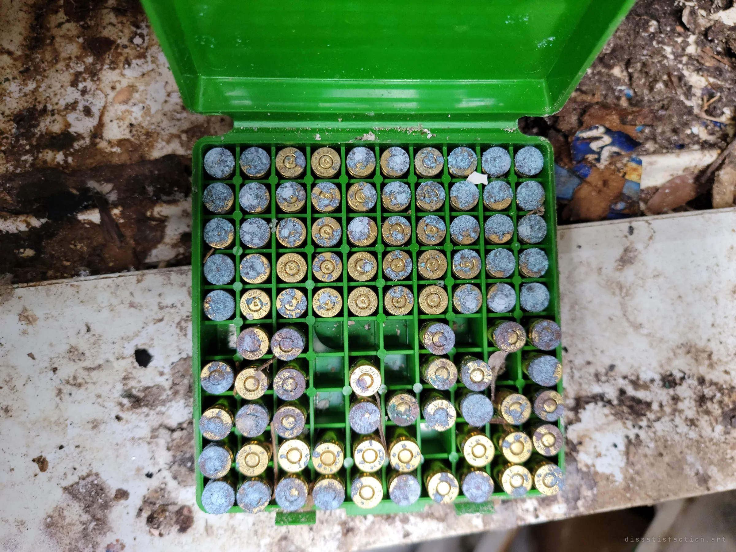 An open green plastic case of pistol cartridges, the upper rows crusted with pale blue corrosion, resting on a stained white surface. Rows of rounds left to oxidise in the damp.