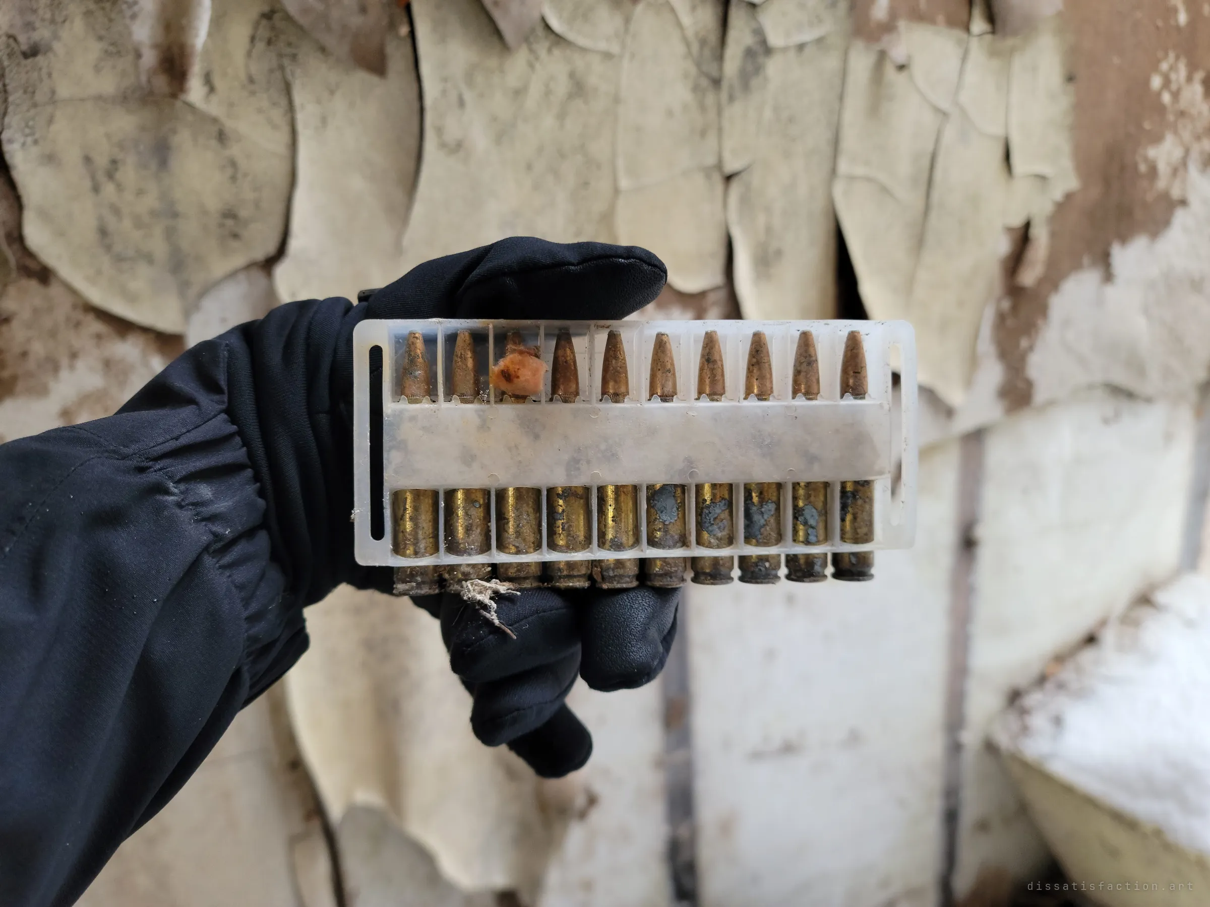 A gloved hand holds up a clear plastic tray of ten rifle cartridges against a wall of curling, peeling paint. The tarnished brass gives a sense of scale to the stockpile.
