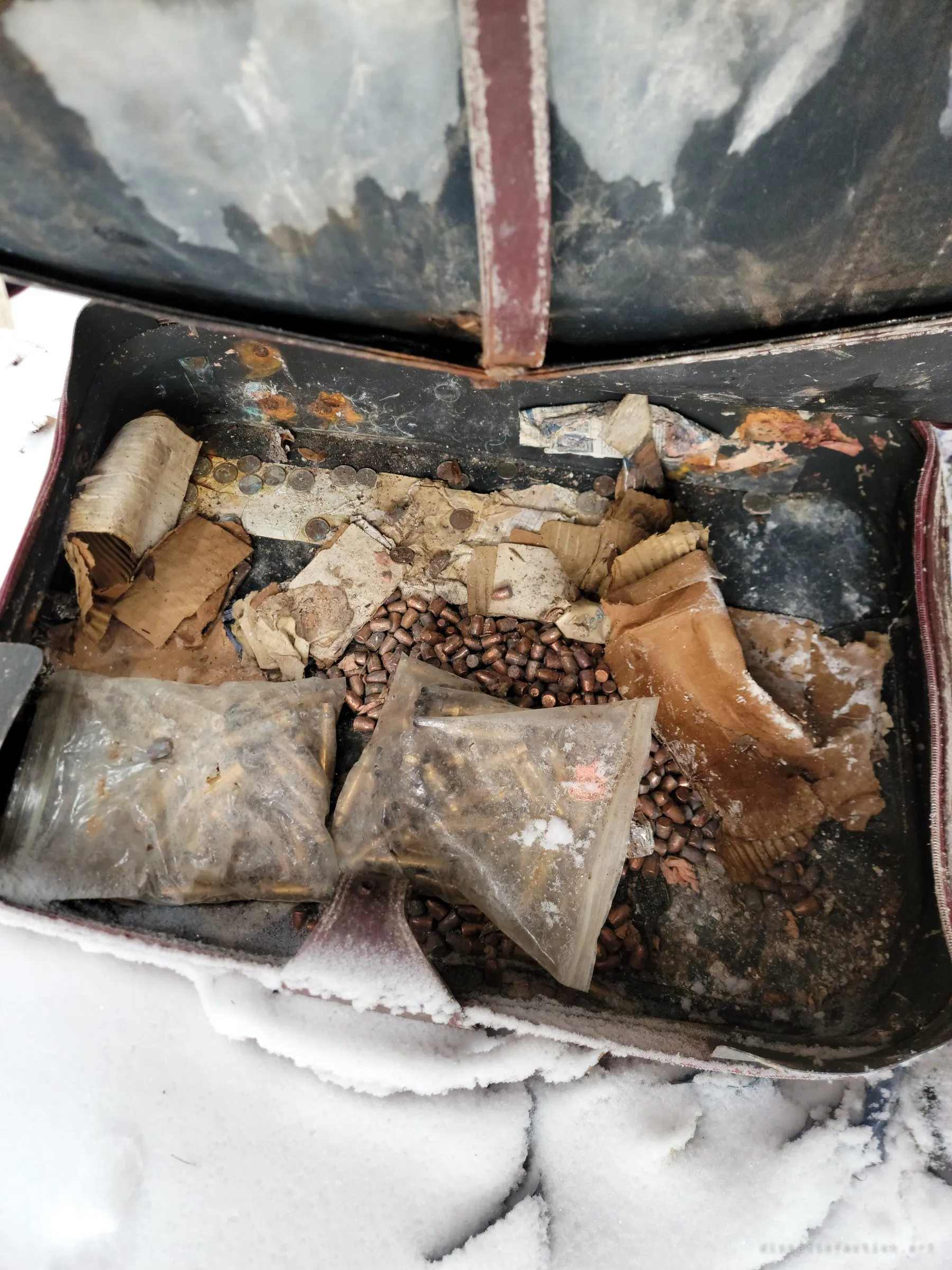 An old leather-strapped case sits open in the snow, filled with copper bullet heads, bags of brass casings, coins and crumbling paper wrappings. Supplies packed away and never used.