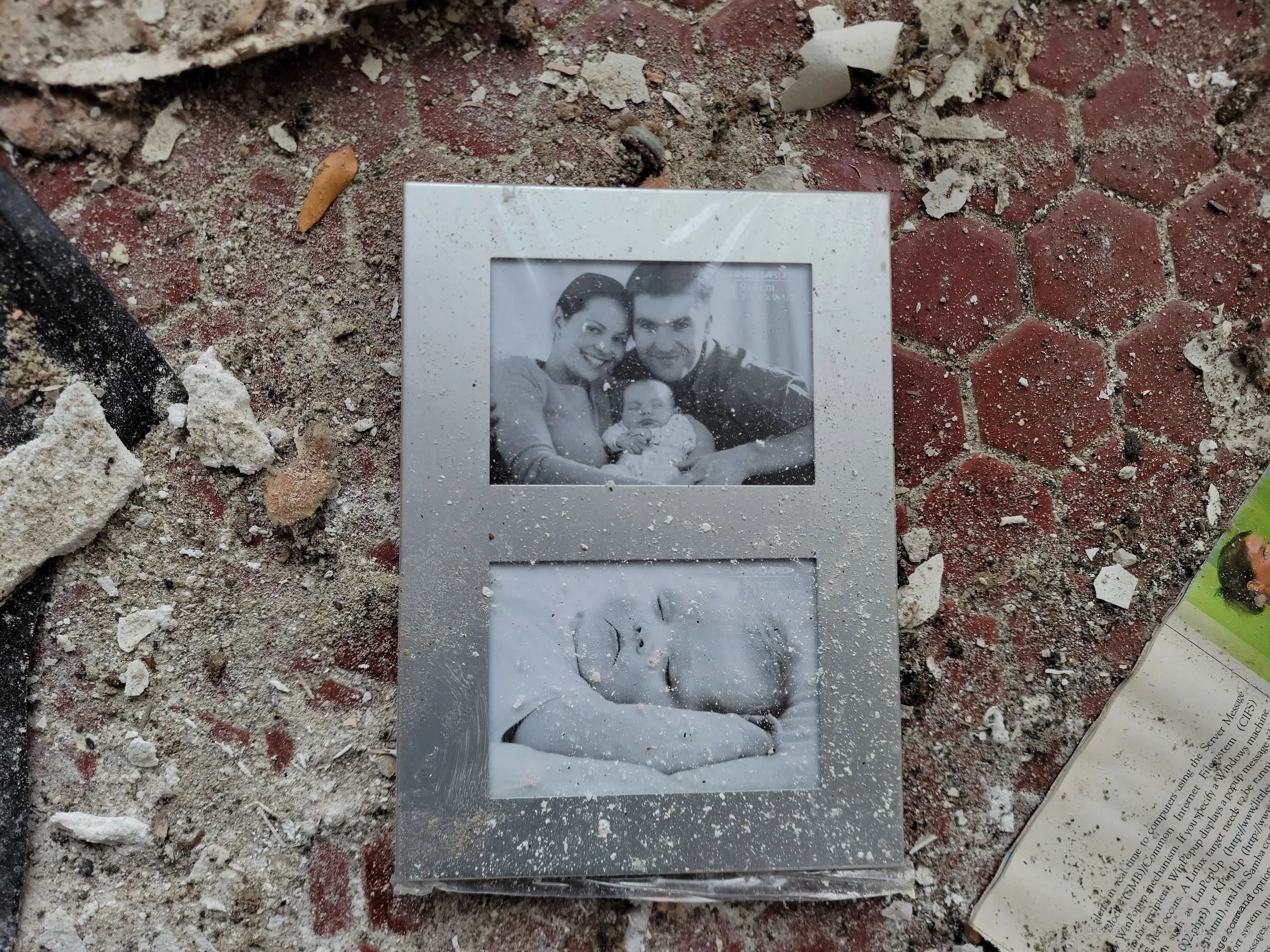 A dusty silver double photo frame lies on a red hexagon tile floor among plaster chunks, still holding its black-and-white pictures of a young family and a sleeping baby. A small domestic trace amid the wreckage.