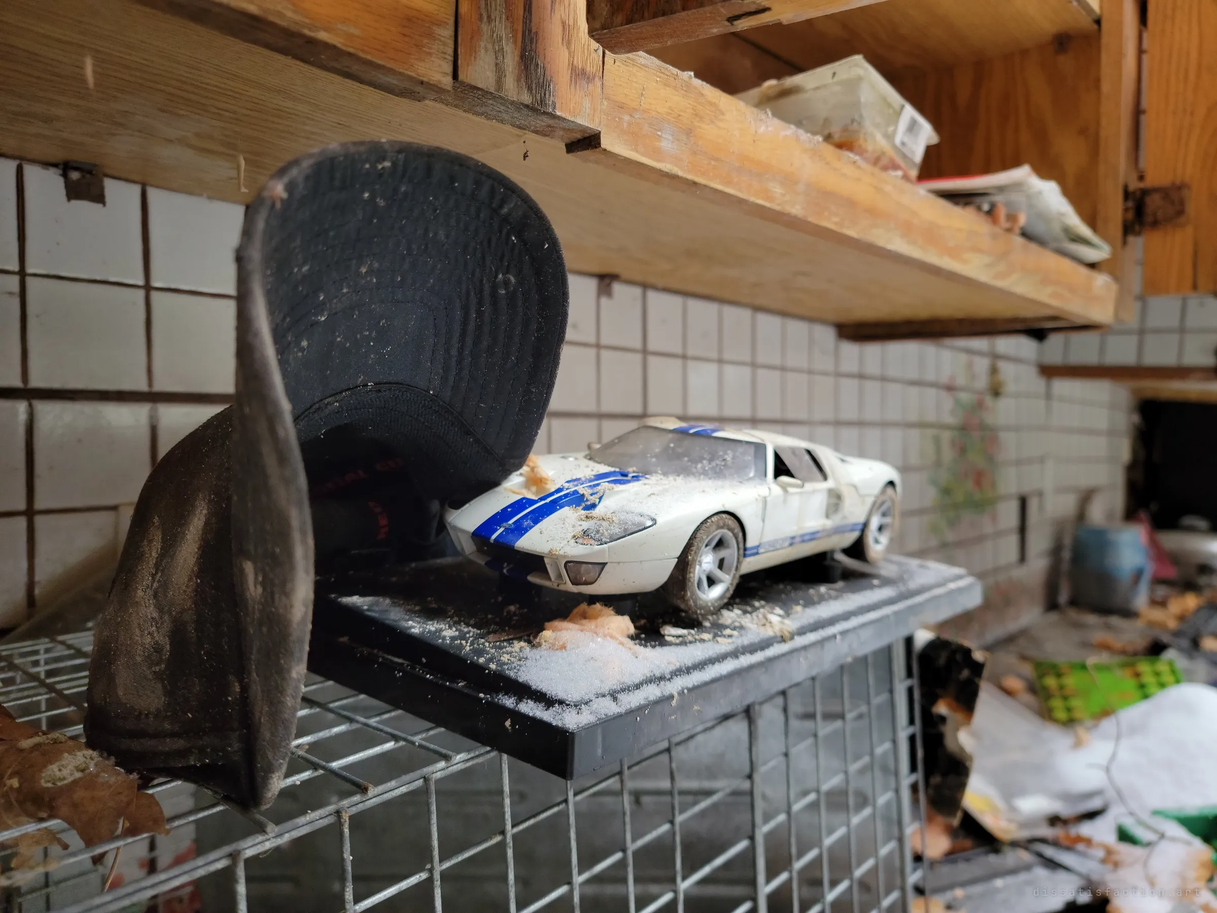 A white model race car with blue stripes sits on a black display base on a wire rack, next to a crumpled cap, below a wooden shelf and a tiled kitchen wall. A collector's piece dusted with snow and insulation.