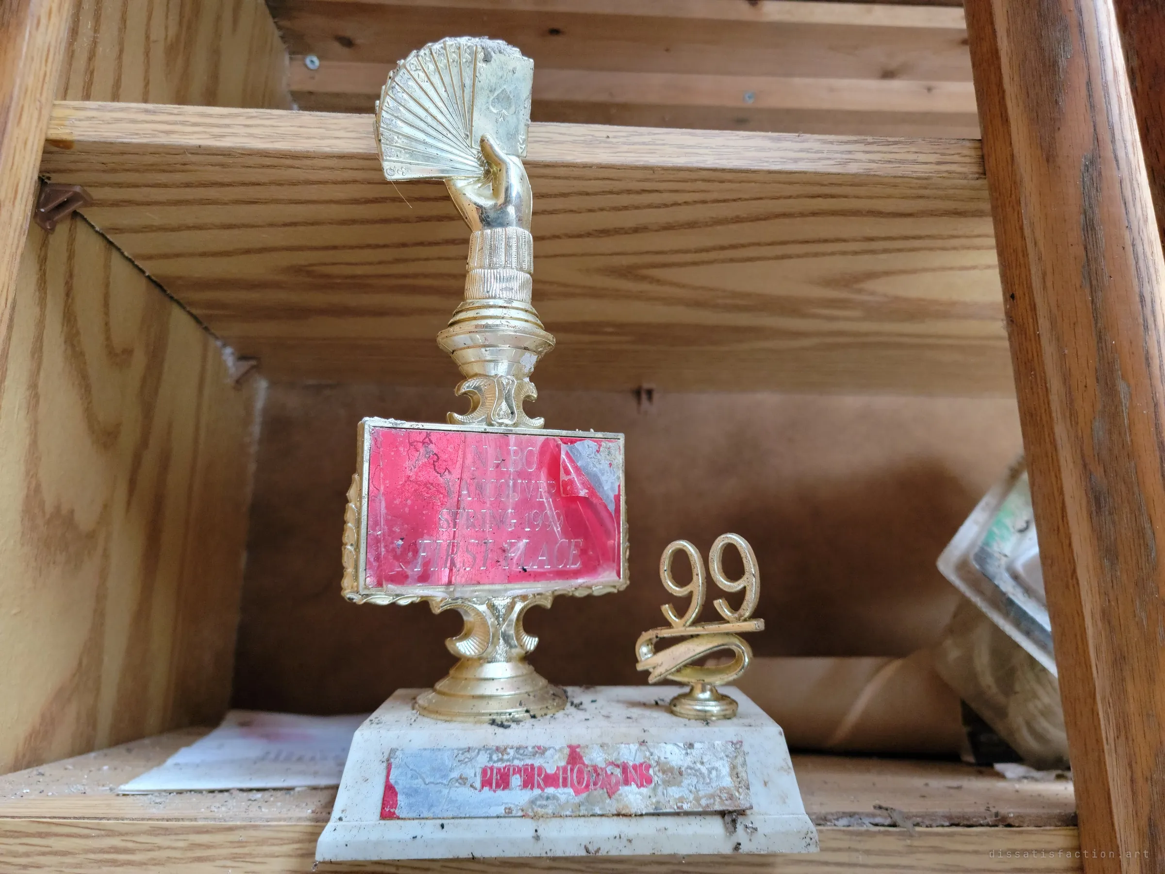 A gold card-playing trophy, a hand fanning a deck of cards, stands on a wooden shelf with a separate year topper beside it. A small prize from a hobby the owner once competed in.