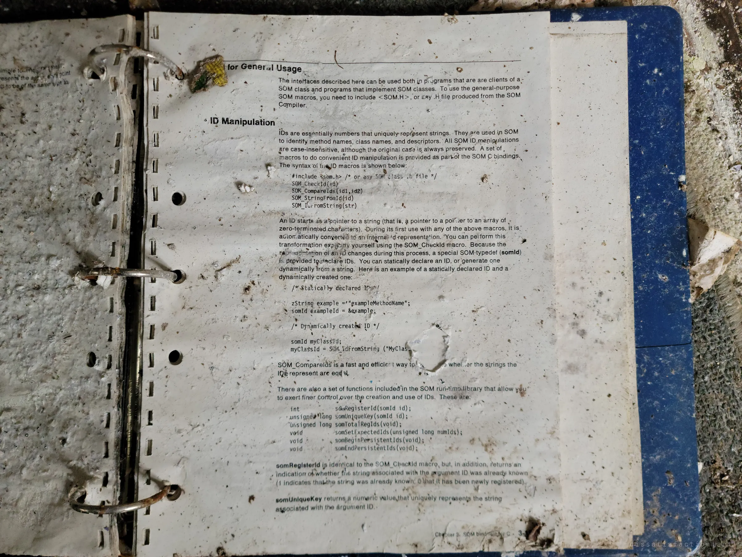 A water-stained ring binder lies open on a page of an old programming manual, its print warped and speckled with debris. Technical reading left behind with everything else.
