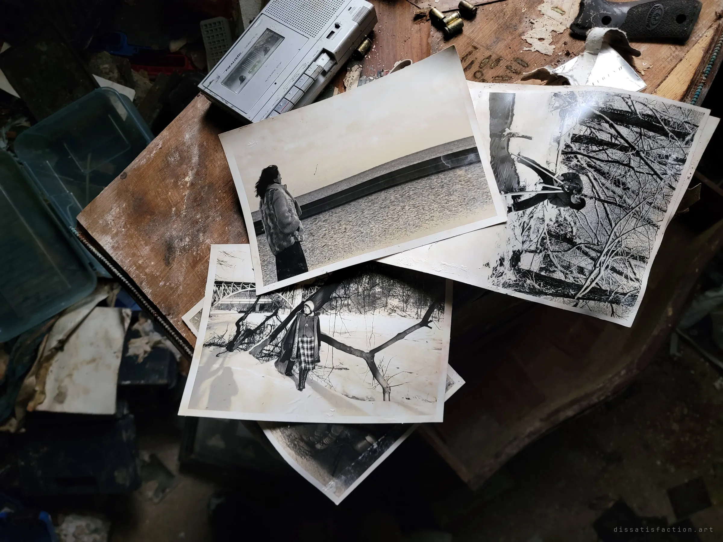 Old black-and-white prints of a woman by the water and in a snowy wood lie on a wooden table with a cassette recorder, loose casings and a pistol grip. Personal memories scattered next to the arsenal.