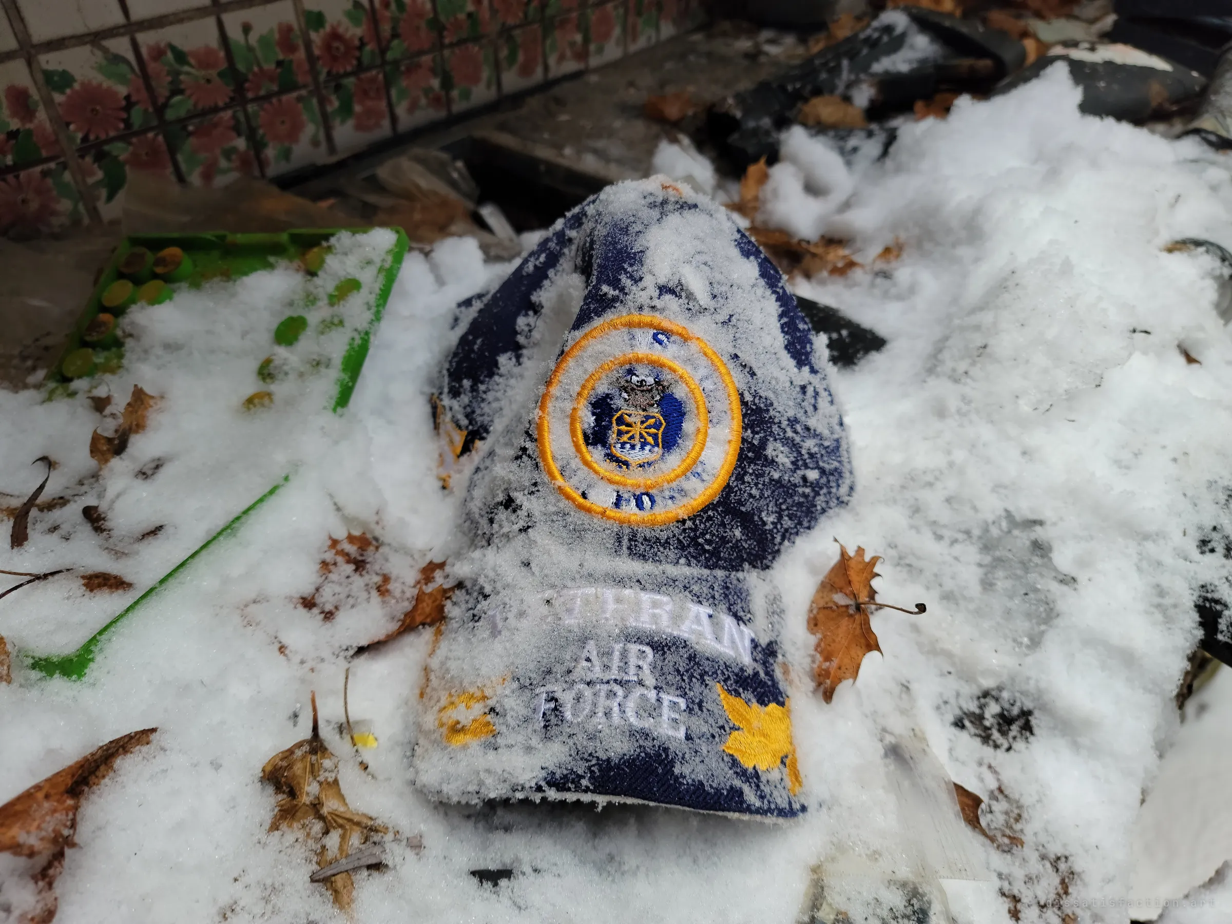 A snow-dusted veteran cap with an embroidered air force crest lies in drifted snow and leaves beside floral tiles, weather already reaching inside the house.
