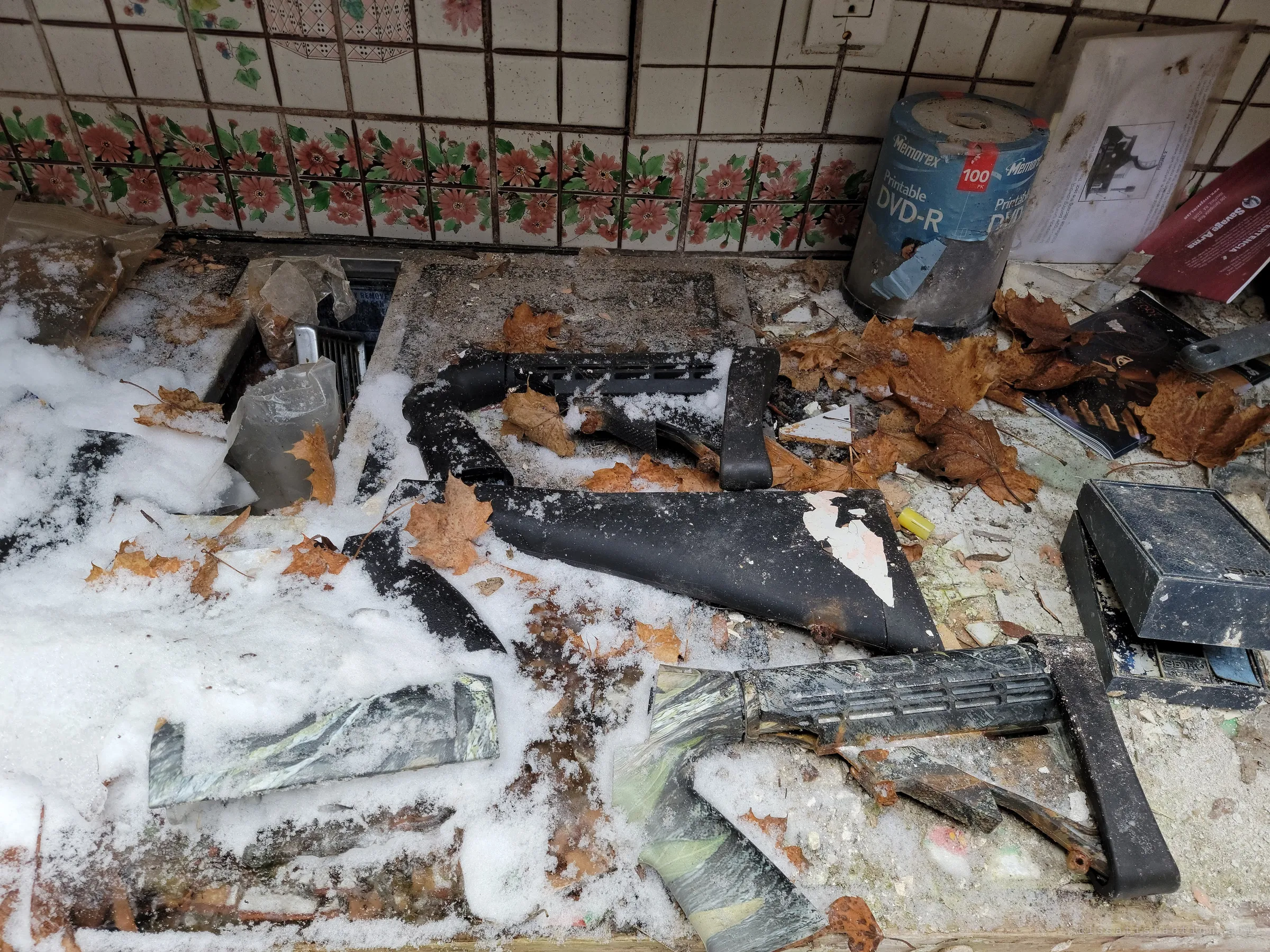 Rifle stocks, some in camouflage finish, lie scattered in snow and leaves on a tiled counter next to a spindle of blank DVDs and firearm manuals, the hobby bench of a prepper left open to the elements.