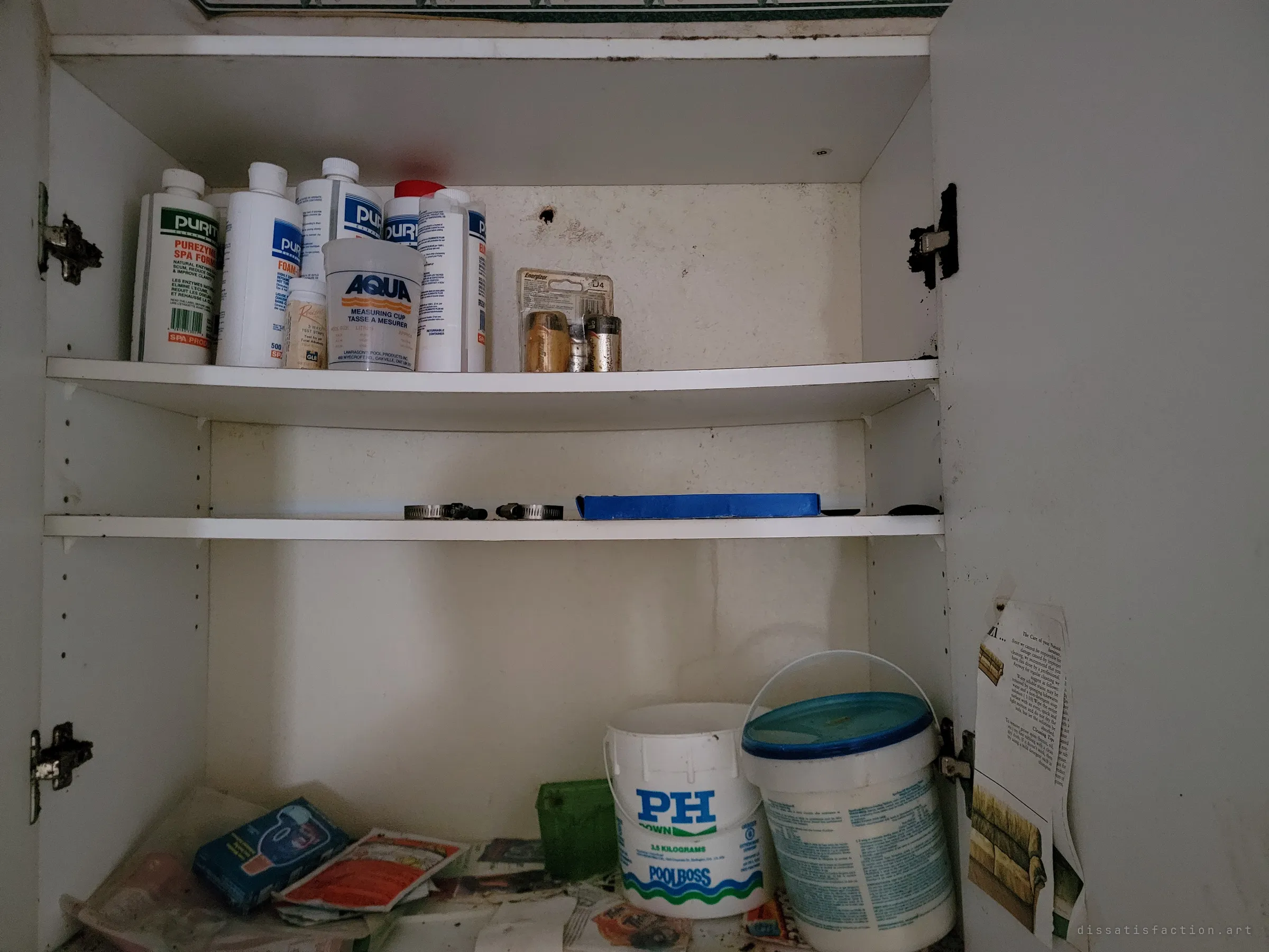 An open cabinet of spa and pool chemicals, batteries and a pH bucket, the ordinary household supplies still lined up on their shelves.