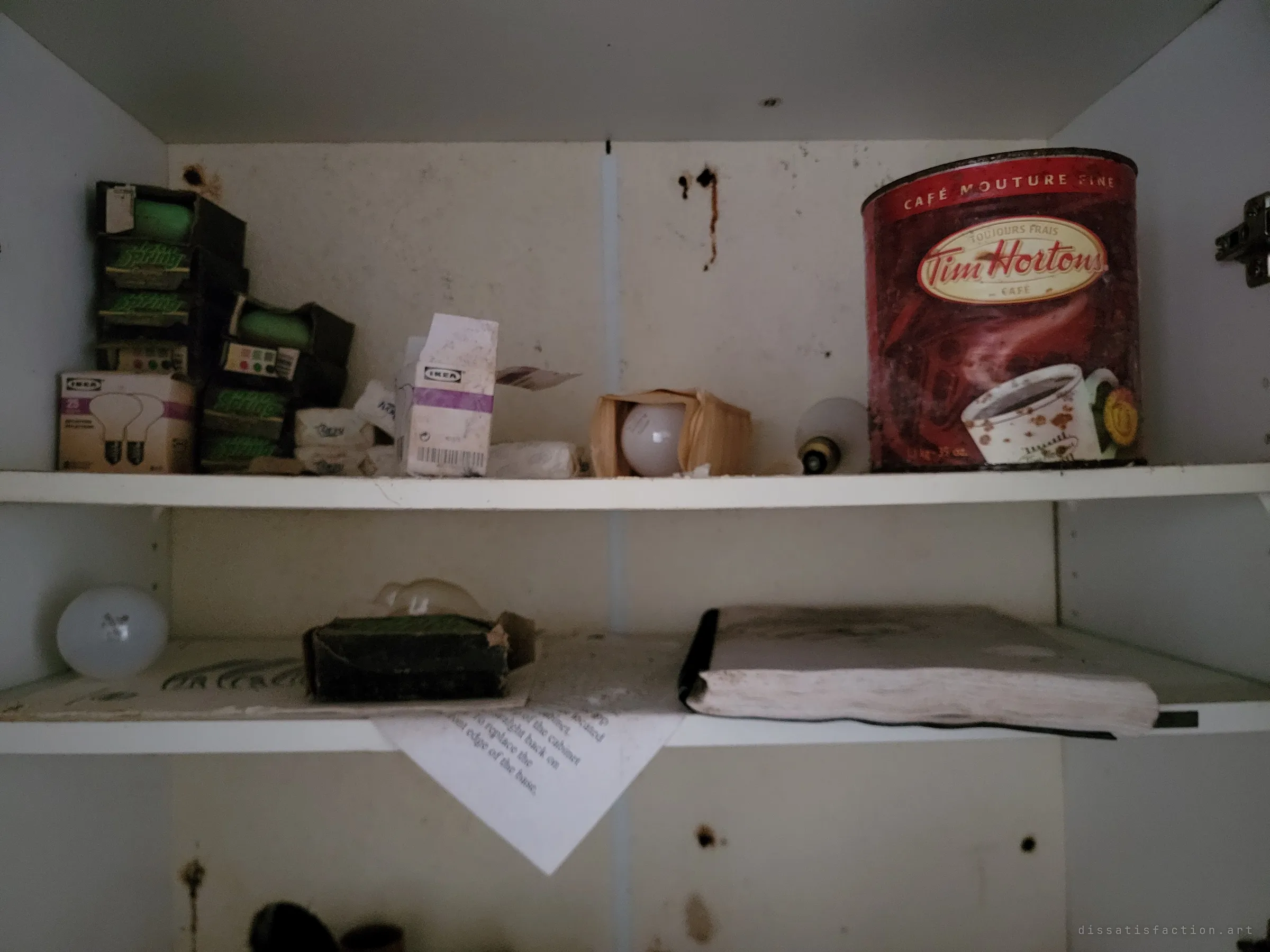 A dim cupboard with boxed light bulbs, a big coffee tin and an old notebook on the shelf below, domestic stock left behind.