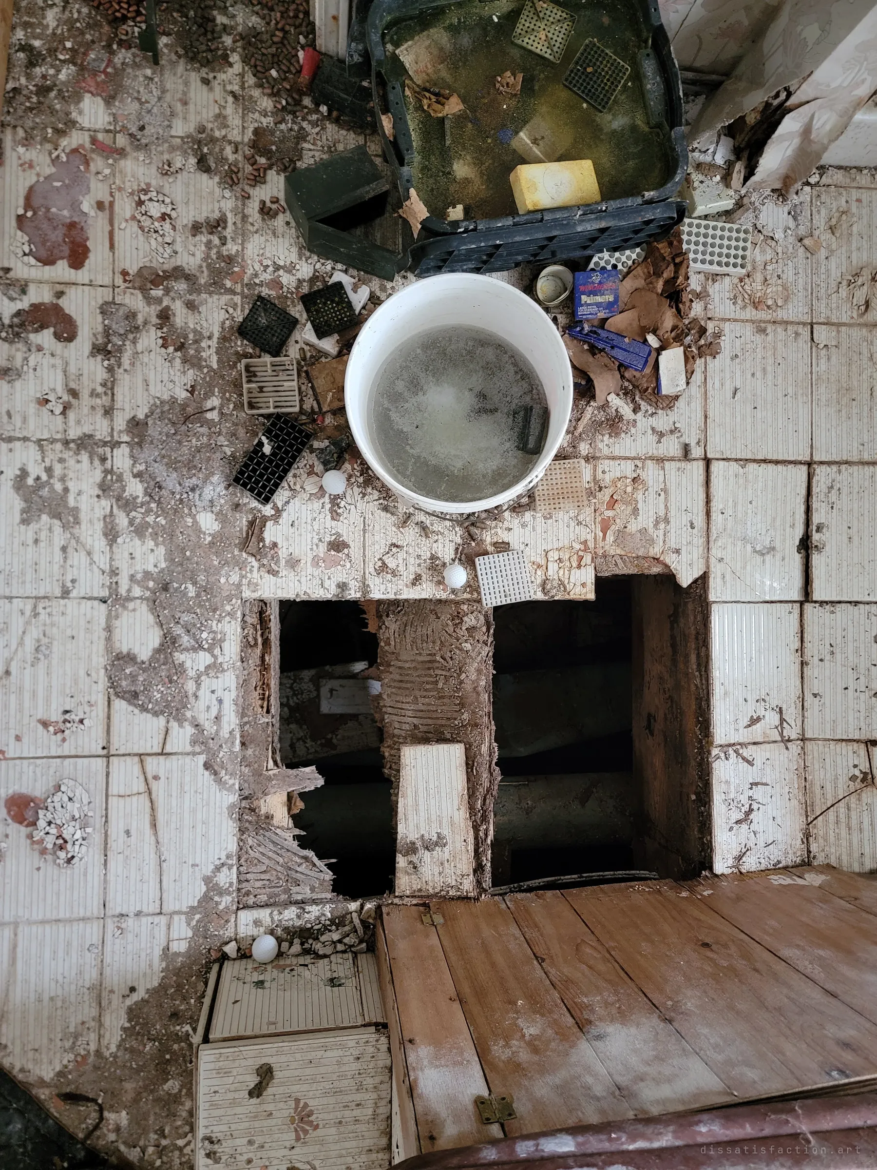 Looking down at a rotted floor with a hole through the tiles, a bucket of grey water and reloading trays scattered with loose bullets and a primer box.