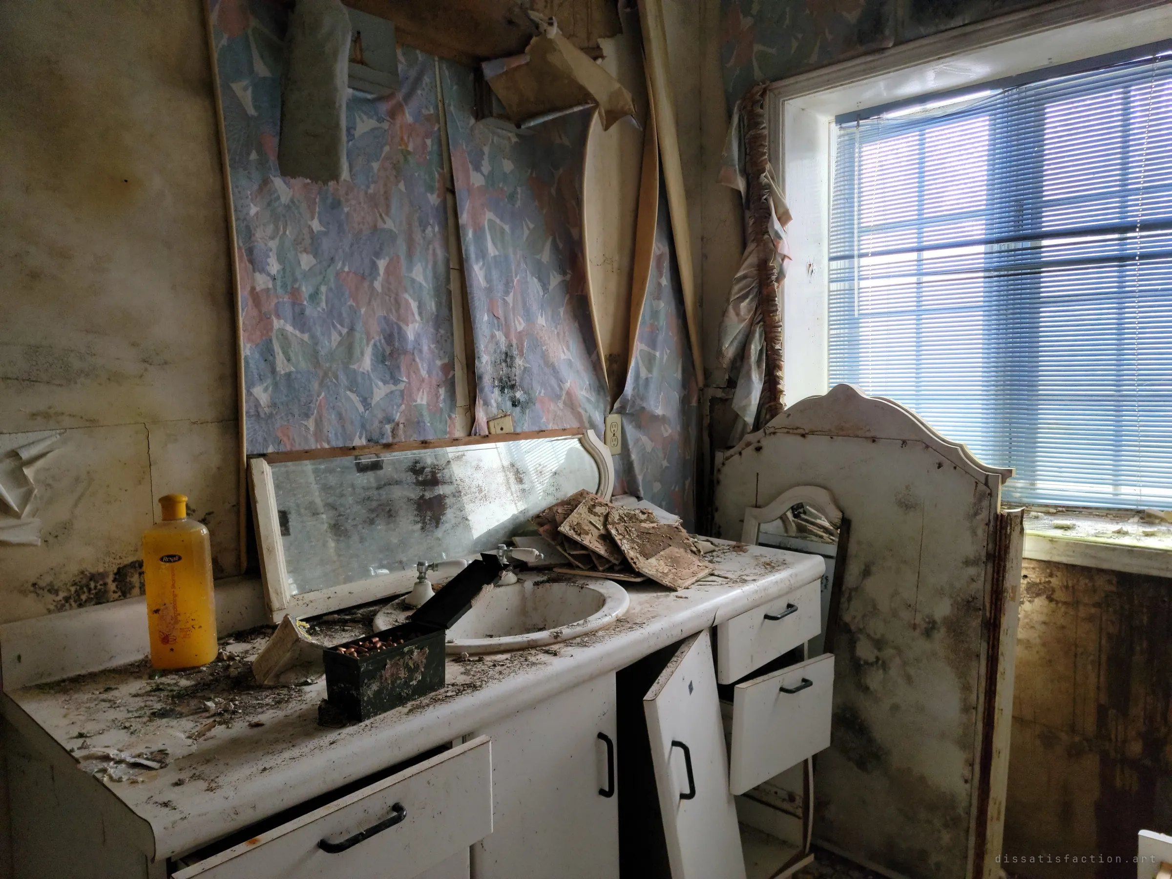 A mould-streaked bathroom vanity with a cracked mirror, fallen ceiling tiles in the sink and an ammo can of cartridges on the counter, lit by blue light through closed blinds.