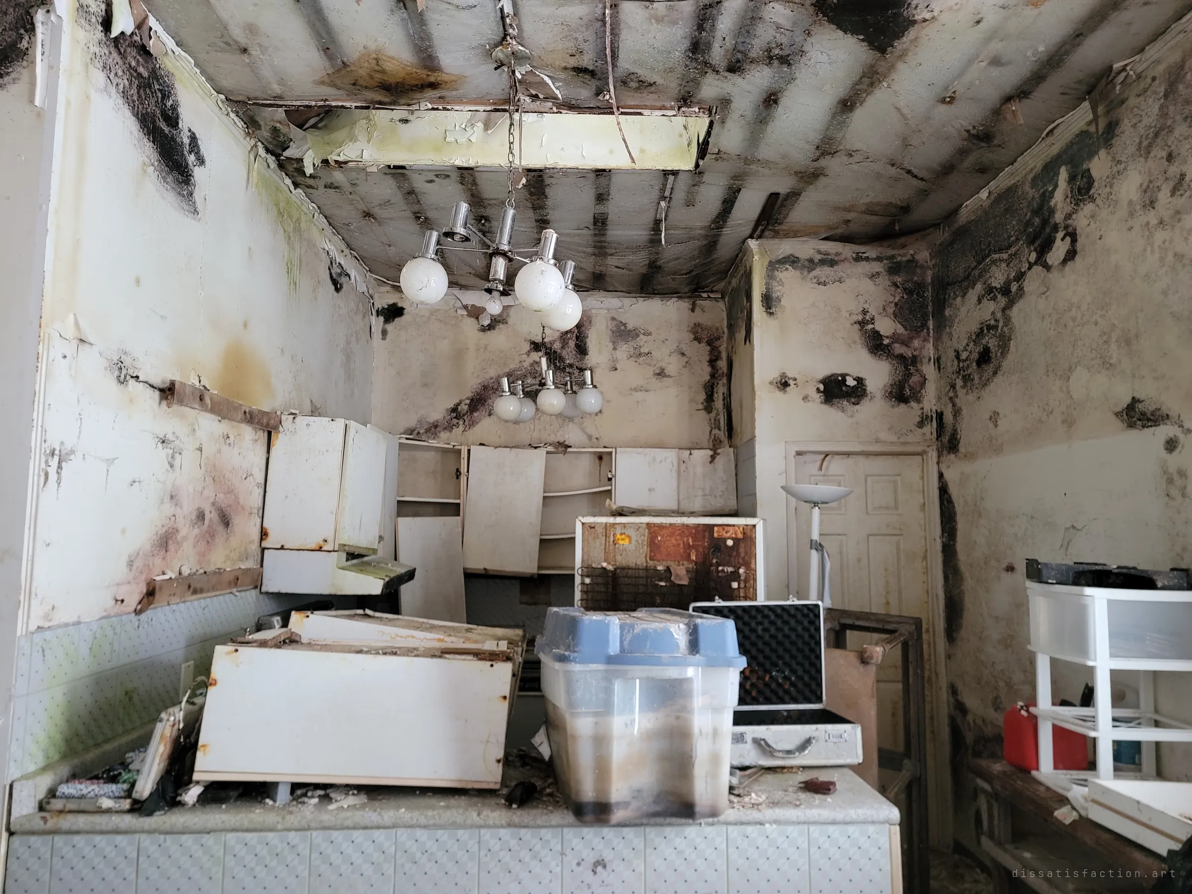 A mould-blackened kitchen with sagging ceiling, twin chrome chandeliers and cabinets torn from the walls, crates and cases piled on the island.