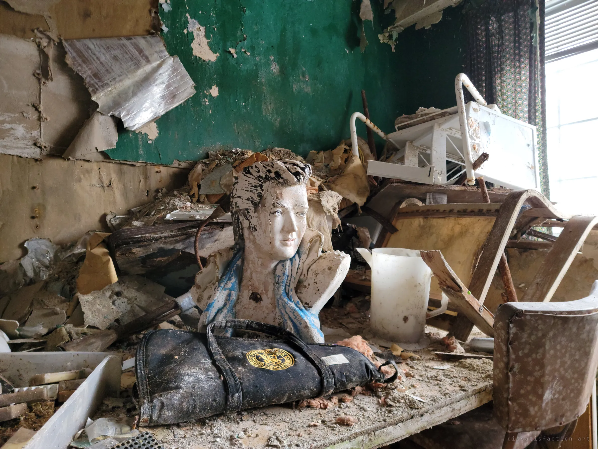 A chipped plaster bust with a blue scarf stands among collapsed debris and a black gun-cleaning roll against a peeling green wall, a strange keepsake in the ruins.