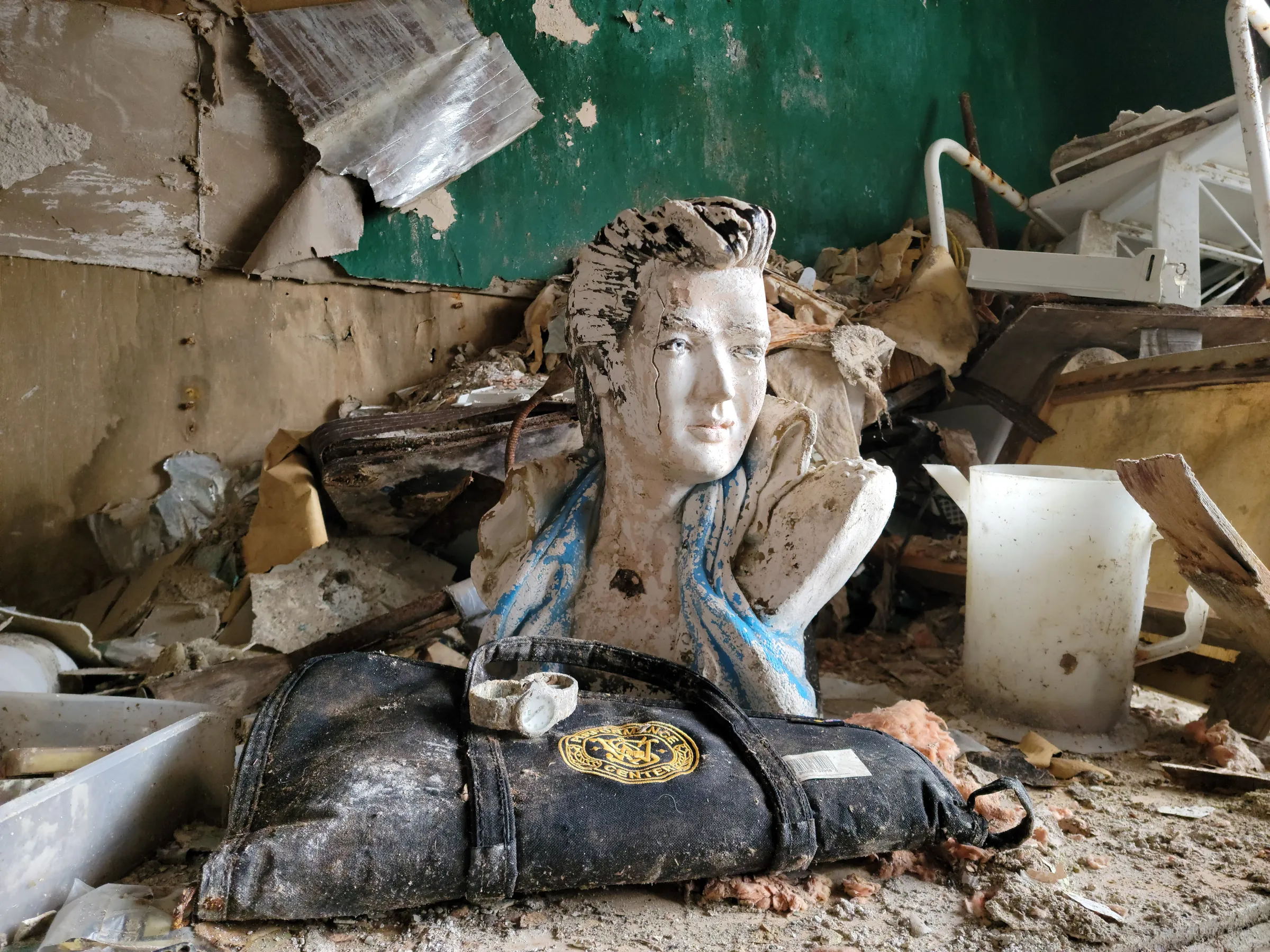 A peeling plaster bust of a pompadoured singer sits in the rubble in front of a torn green wall, with a mud-caked wristwatch resting on a black embroidered rifle case. Kitsch, a weapon case and debris piled together sum up the prepper's house.