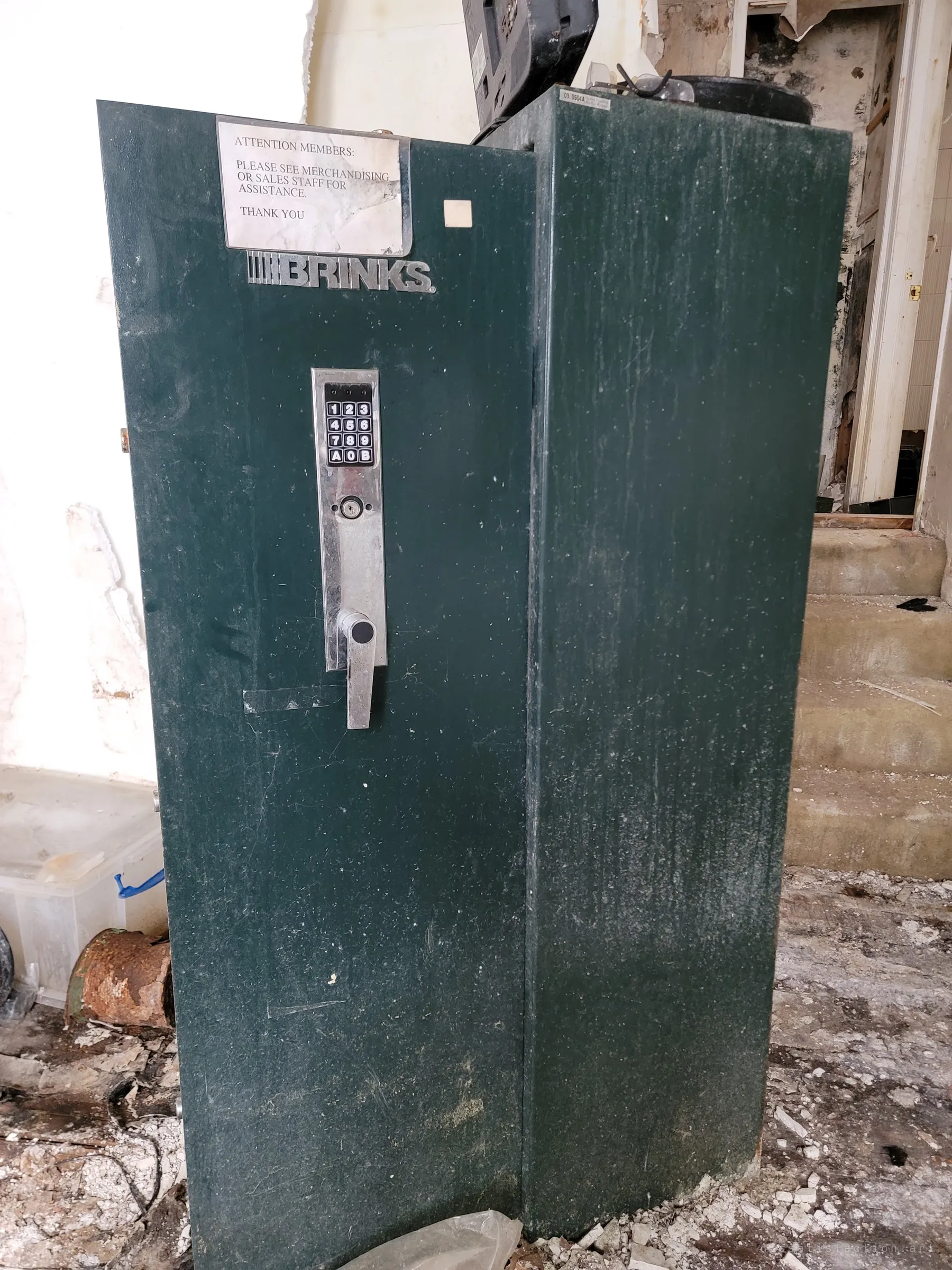 A tall green gun safe with a keypad lock and a faded shop notice taped to its door stands by a set of worn concrete steps. The store-issued safe hints at the arsenal once kept here.