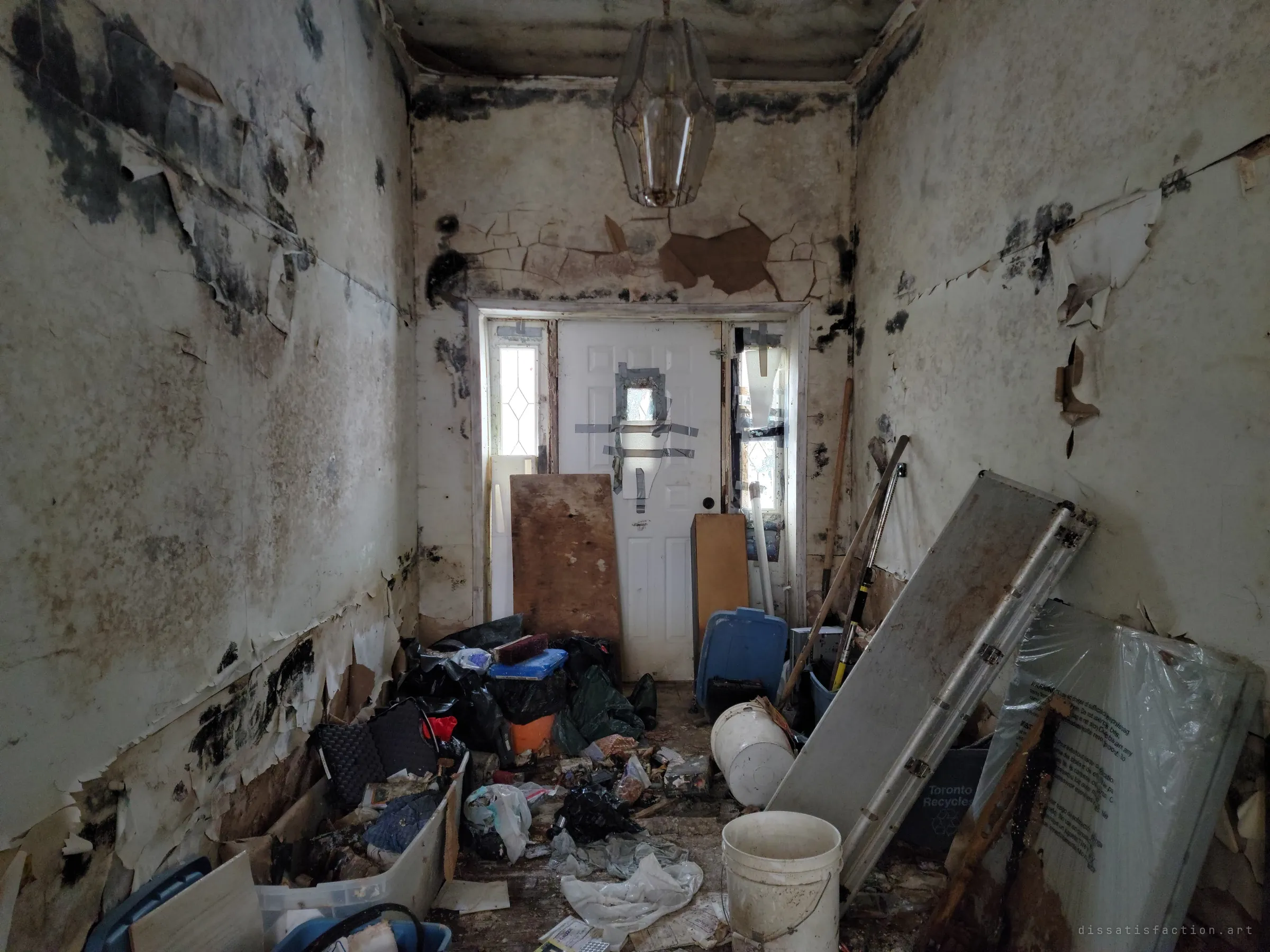 A narrow entry hall with mould-blackened, peeling walls runs to a taped-up front door, its floor buried in bags, buckets and boxes. A glass lantern still hangs above the mess.