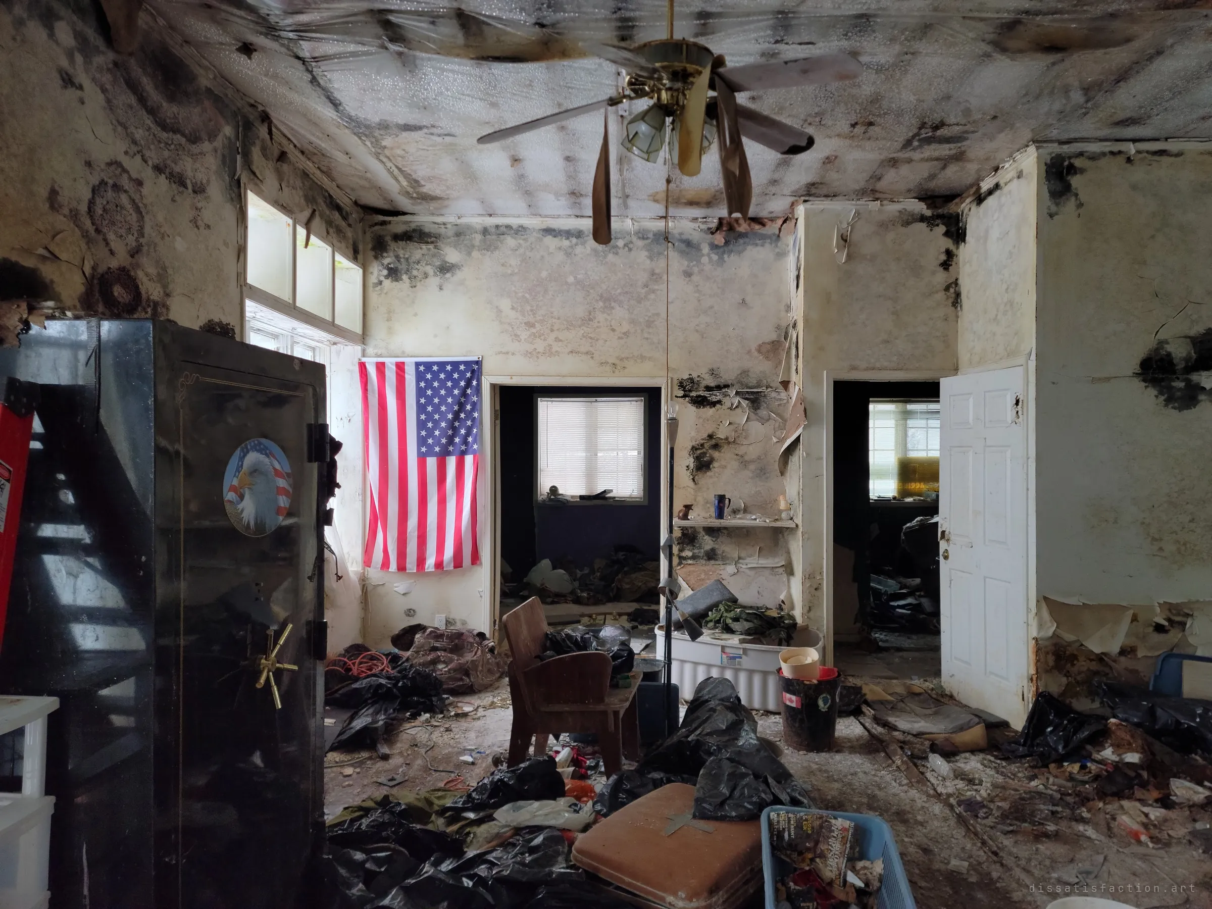 A mould-streaked living room with a US flag on the wall, an eagle-decorated gun safe to the left and a drooping ceiling fan above piles of black bags and camouflage gear. It is the album's title shot of patriotic prepper decor left to rot.