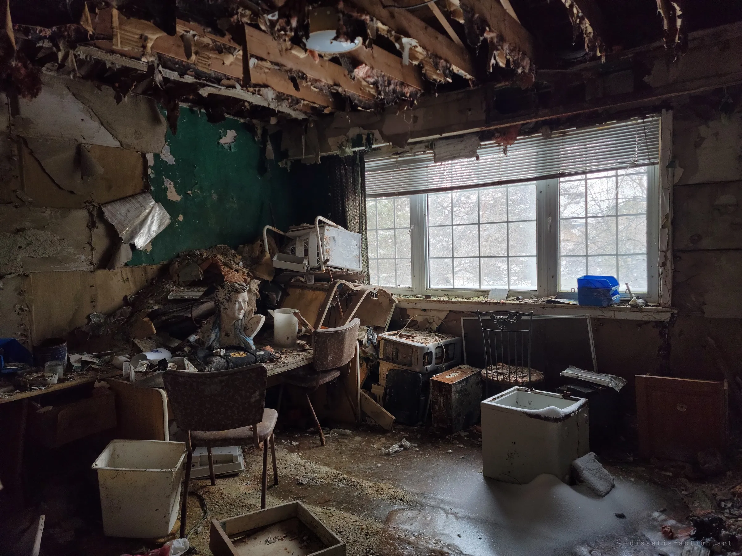 A room whose ceiling has fallen in, with the bust and clutter piled on a desk under a wide window of bare winter trees and snow drifting across the floor. It shows how open to the weather the house has become.