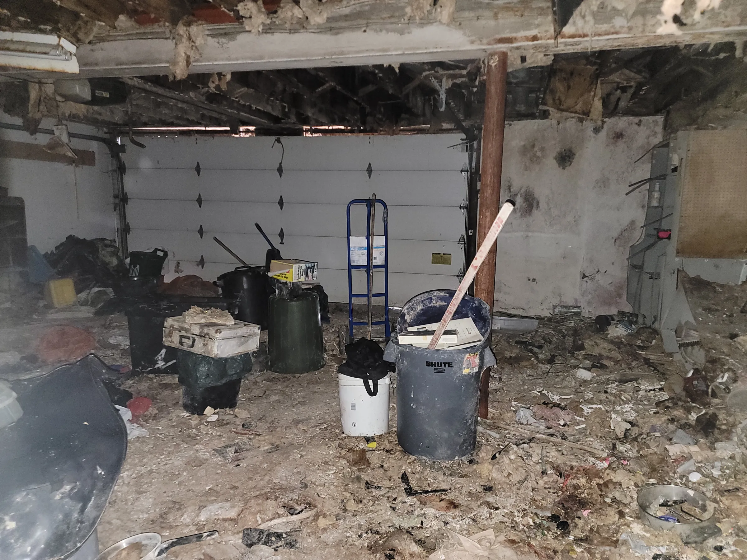 A flash-lit garage with a gutted ceiling, a white roll-up door, a hand truck and trash cans standing in a floor of fallen insulation. The collapse overhead shows how far the decay has spread.