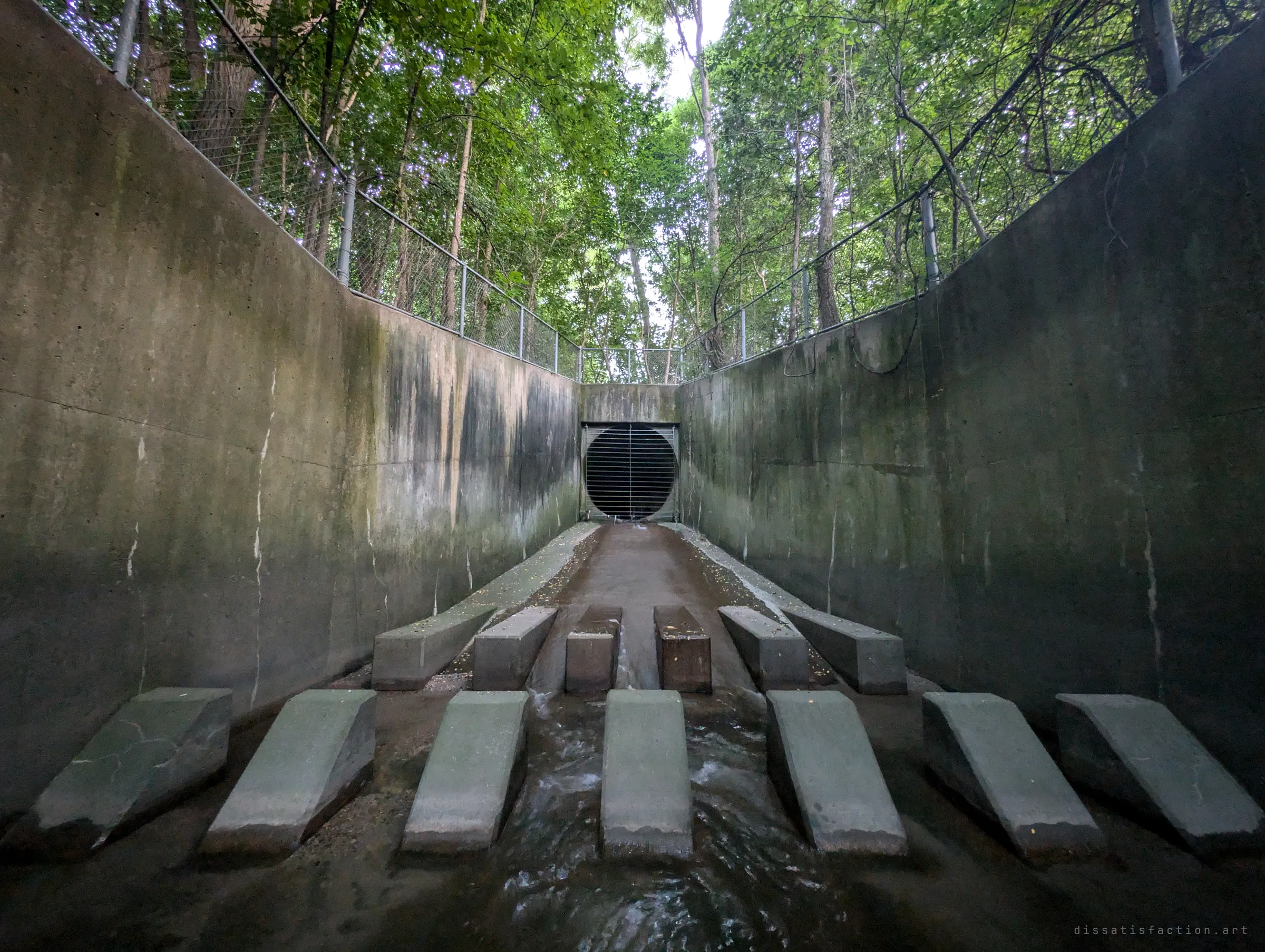 Ultra-wide view down the outfall channel, water rushing between rows of angled concrete baffle blocks toward the barred circular portal. The blocks break the flow before it leaves the drain.