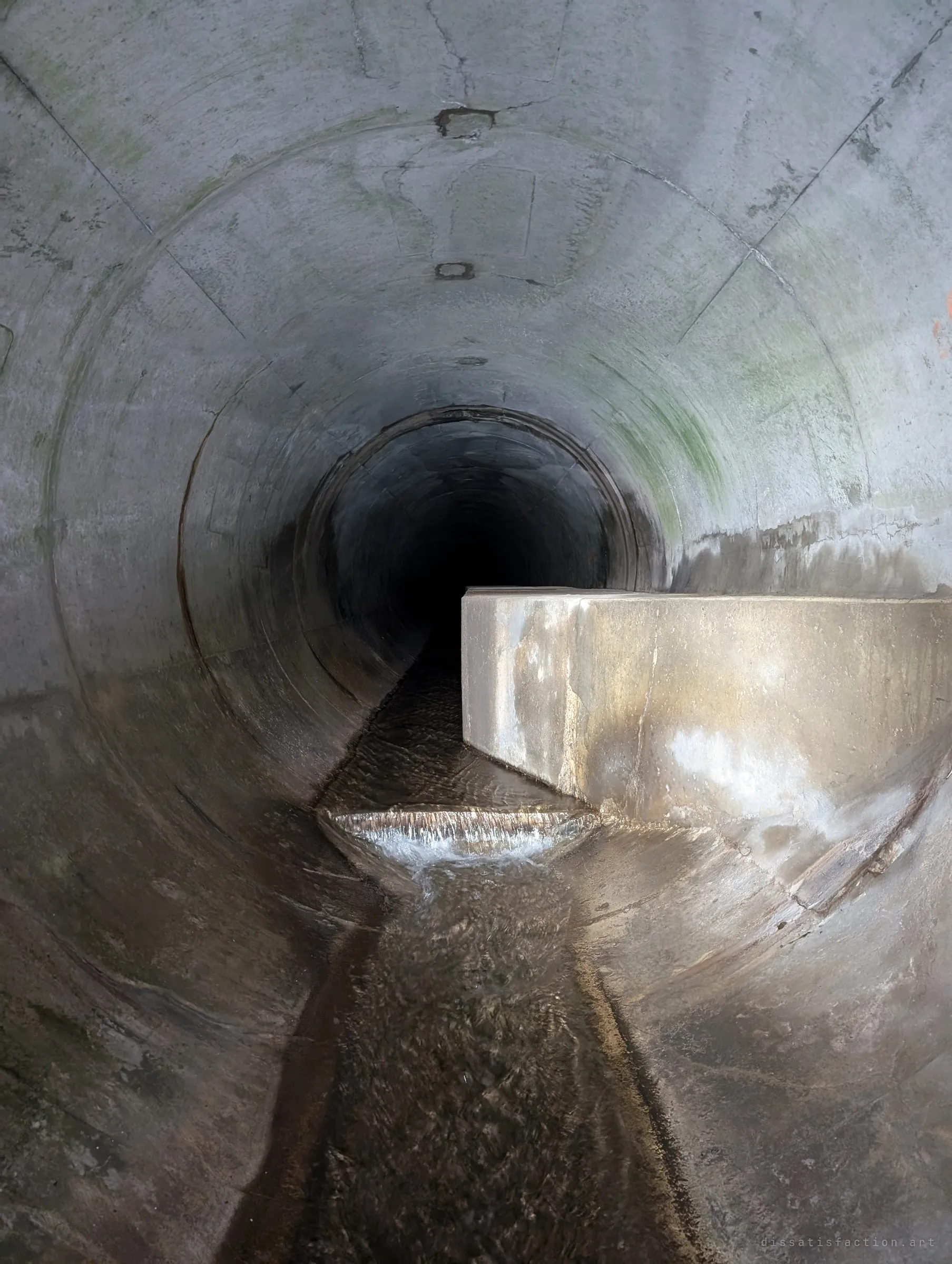 Inside a round concrete pipe, a boxed-in sanitary line runs along one side while a thin sheet of water spills over a small step in the channel. Two systems sharing one tunnel.