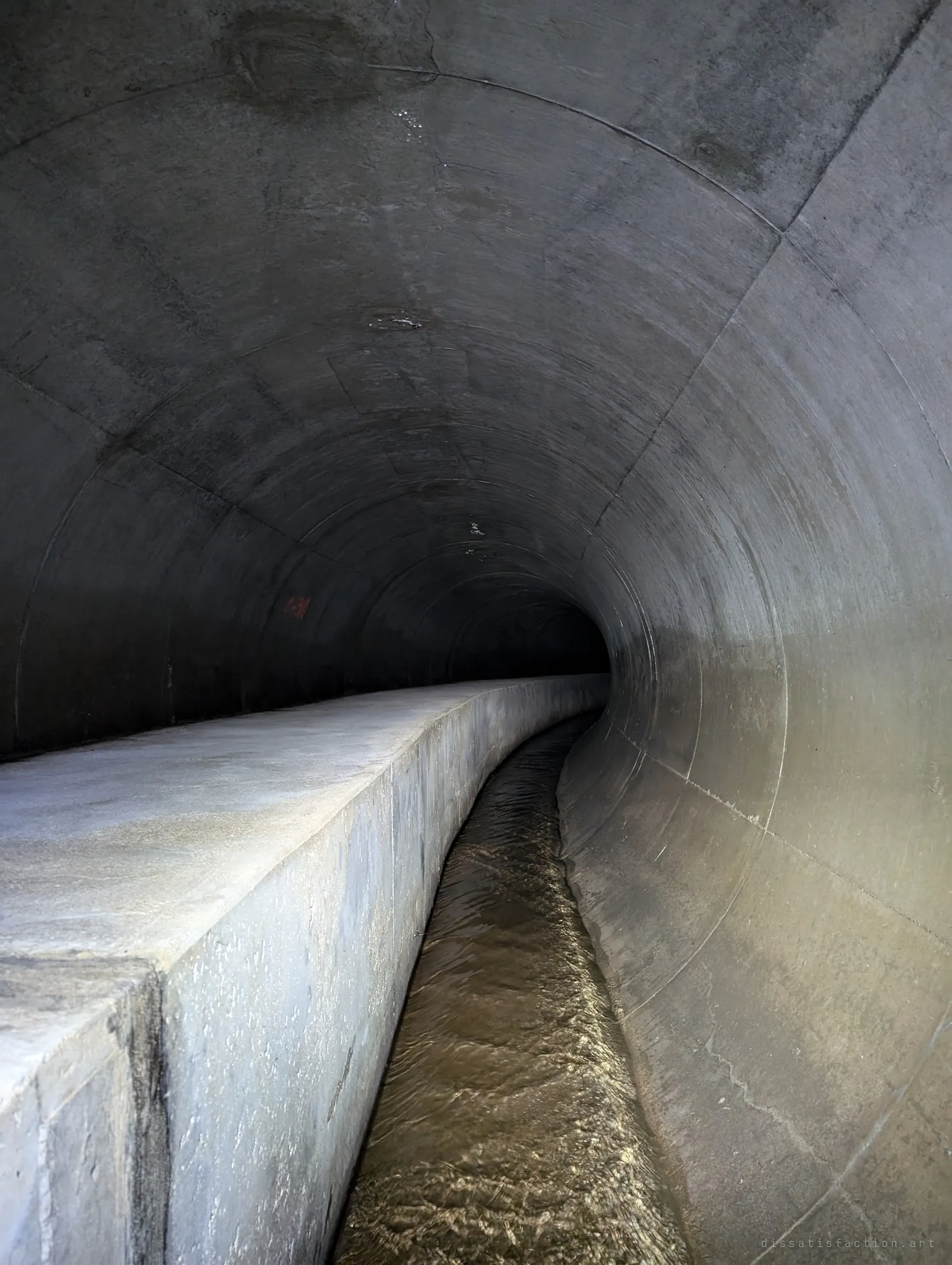 The round tunnel curves gently away into darkness, the concrete sanitary casing on the left and a narrow flowing channel on the right. The bend hides whatever comes next.