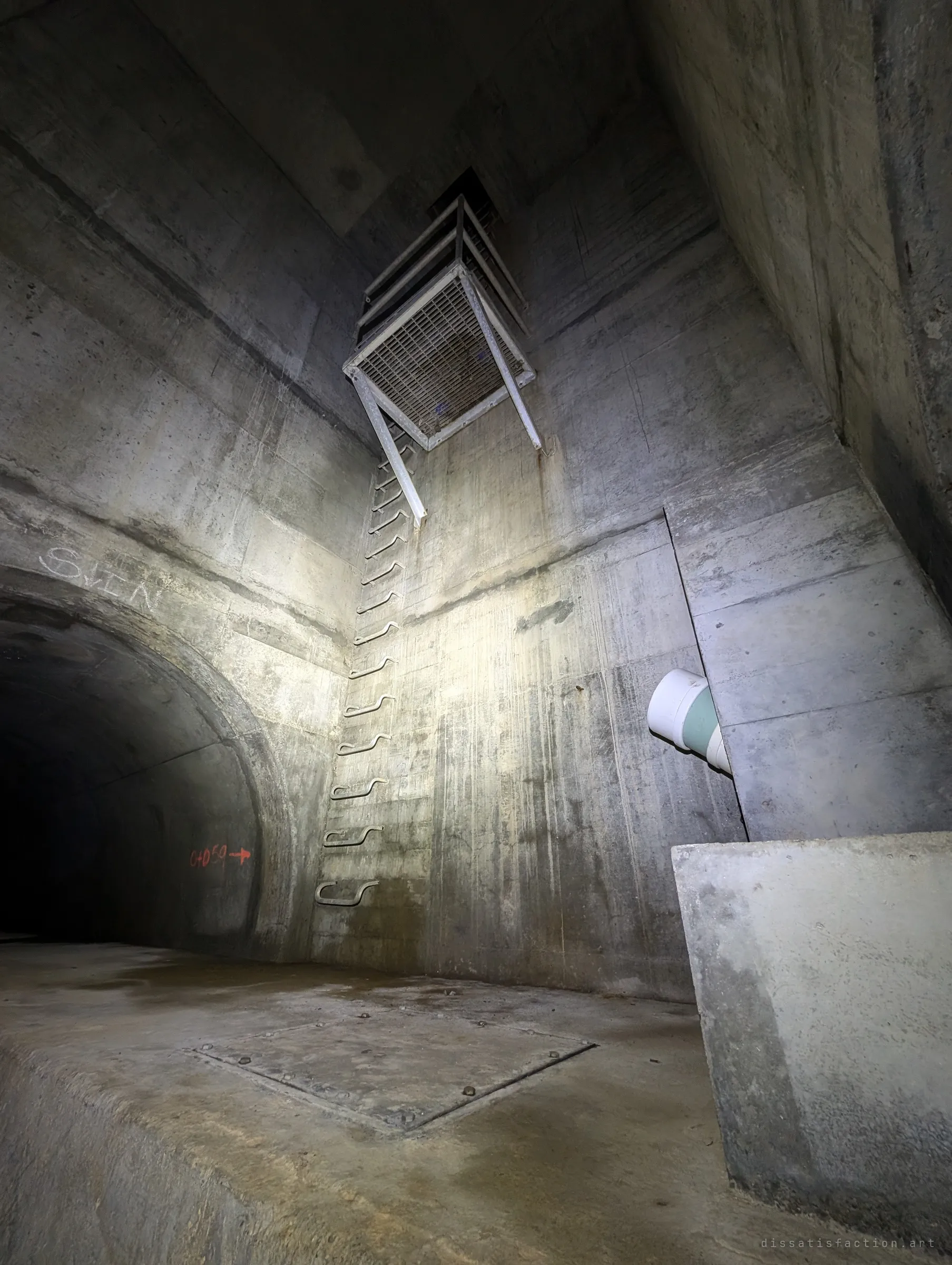 A tall concrete manhole chamber lit from below, a ladder of step irons climbing to a hanging steel landing far overhead, a chalk tag and survey mark by the tunnel mouth. It shows how deep the drain runs.