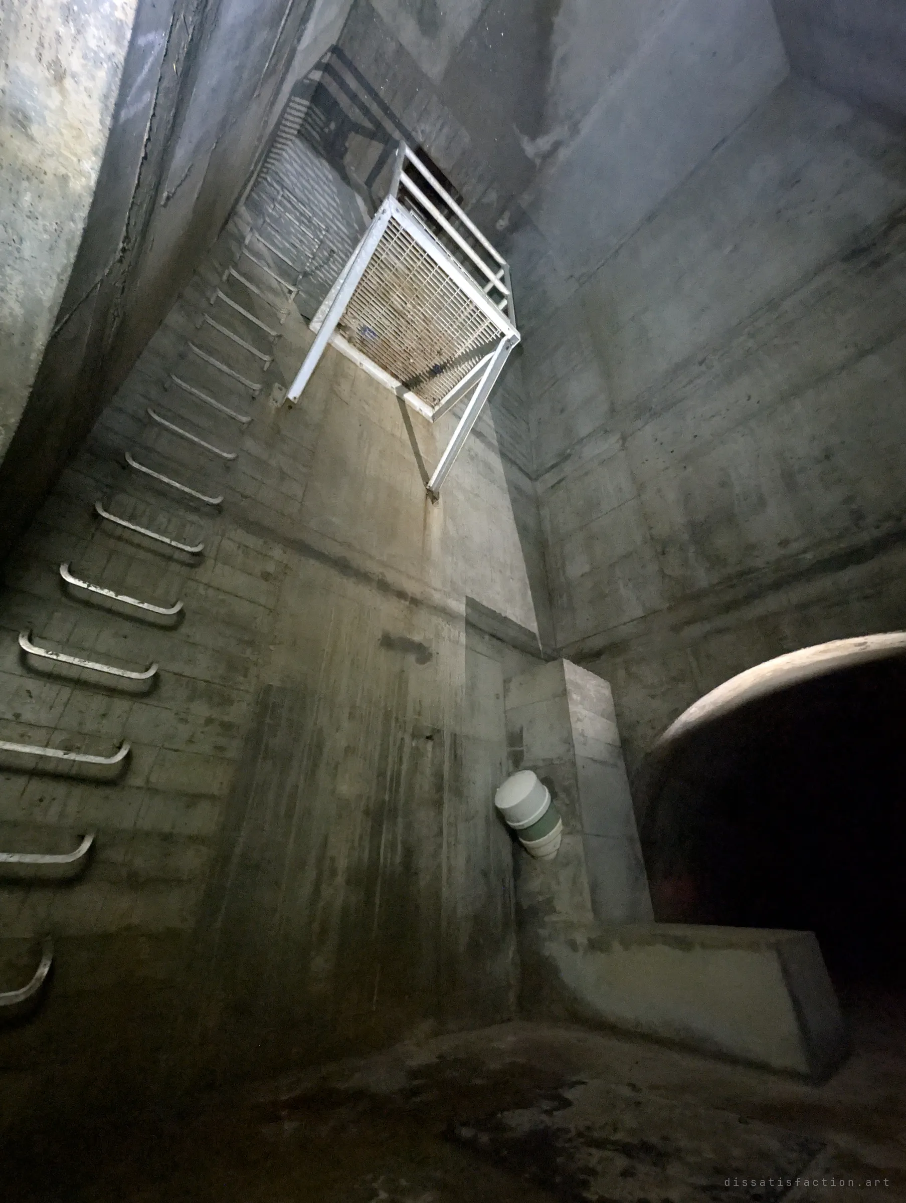 Looking straight up the same chamber: step irons spiral up the wall to a grated steel landing hung in the shaft, with the dark tunnel mouth below right. The vertical scale of the chamber in one frame.