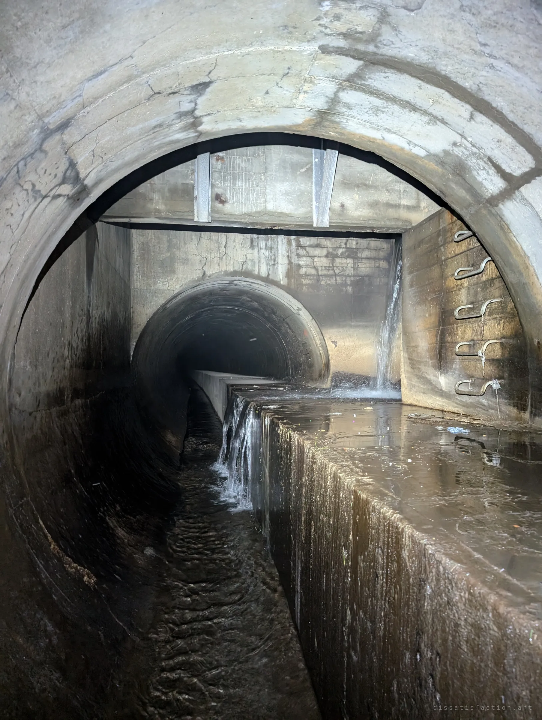 Through a round pipe mouth, water pours from a side inlet into a junction chamber and over a concrete ledge into the lower channel, step irons on the right wall. A spot where you cannot stay dry.