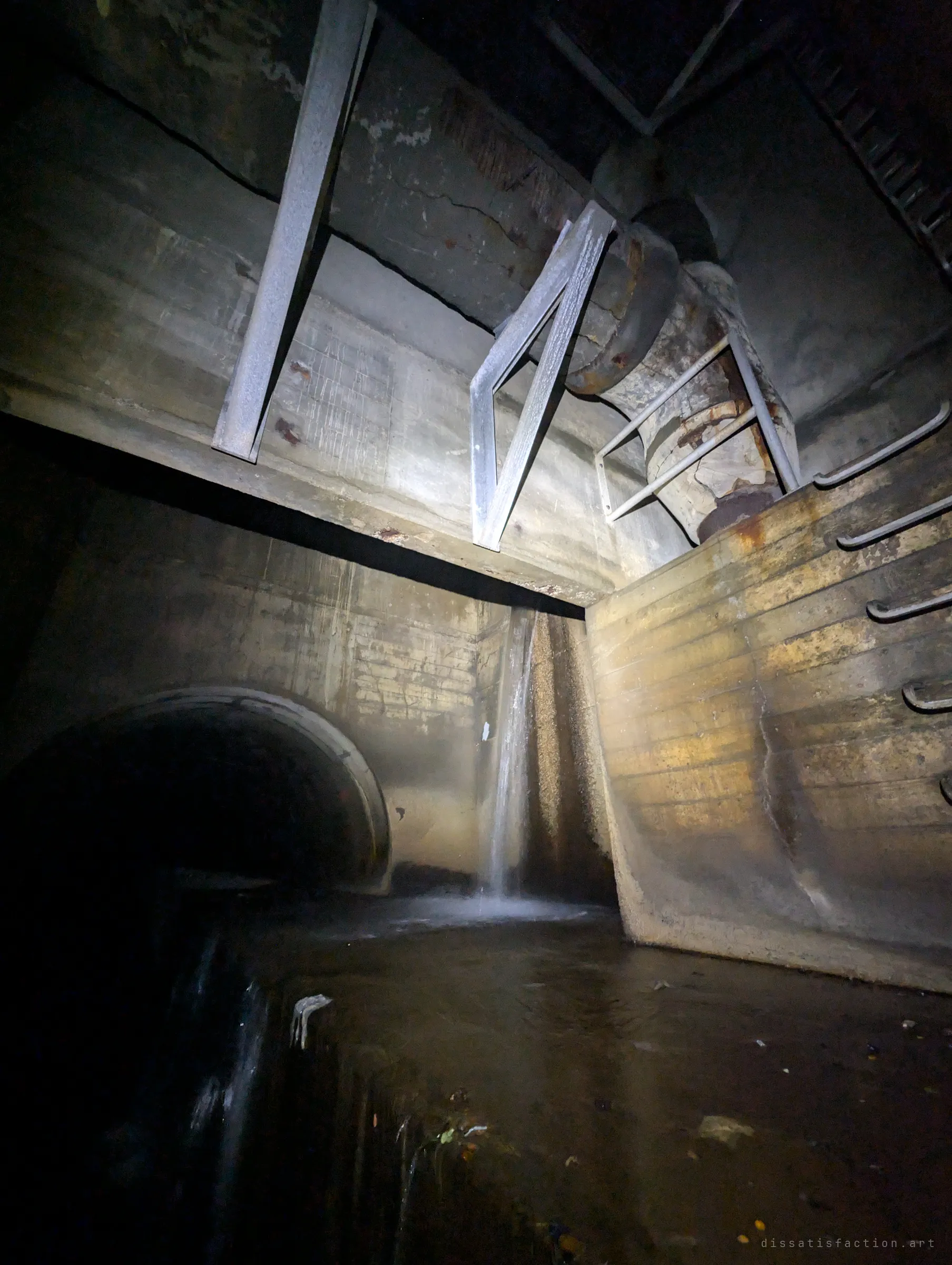 Looking up in the junction chamber: steel braces and a rusted pipe hang overhead while a thin waterfall drops beside a tunnel mouth into dark water. Rust, concrete and falling water in the dark.