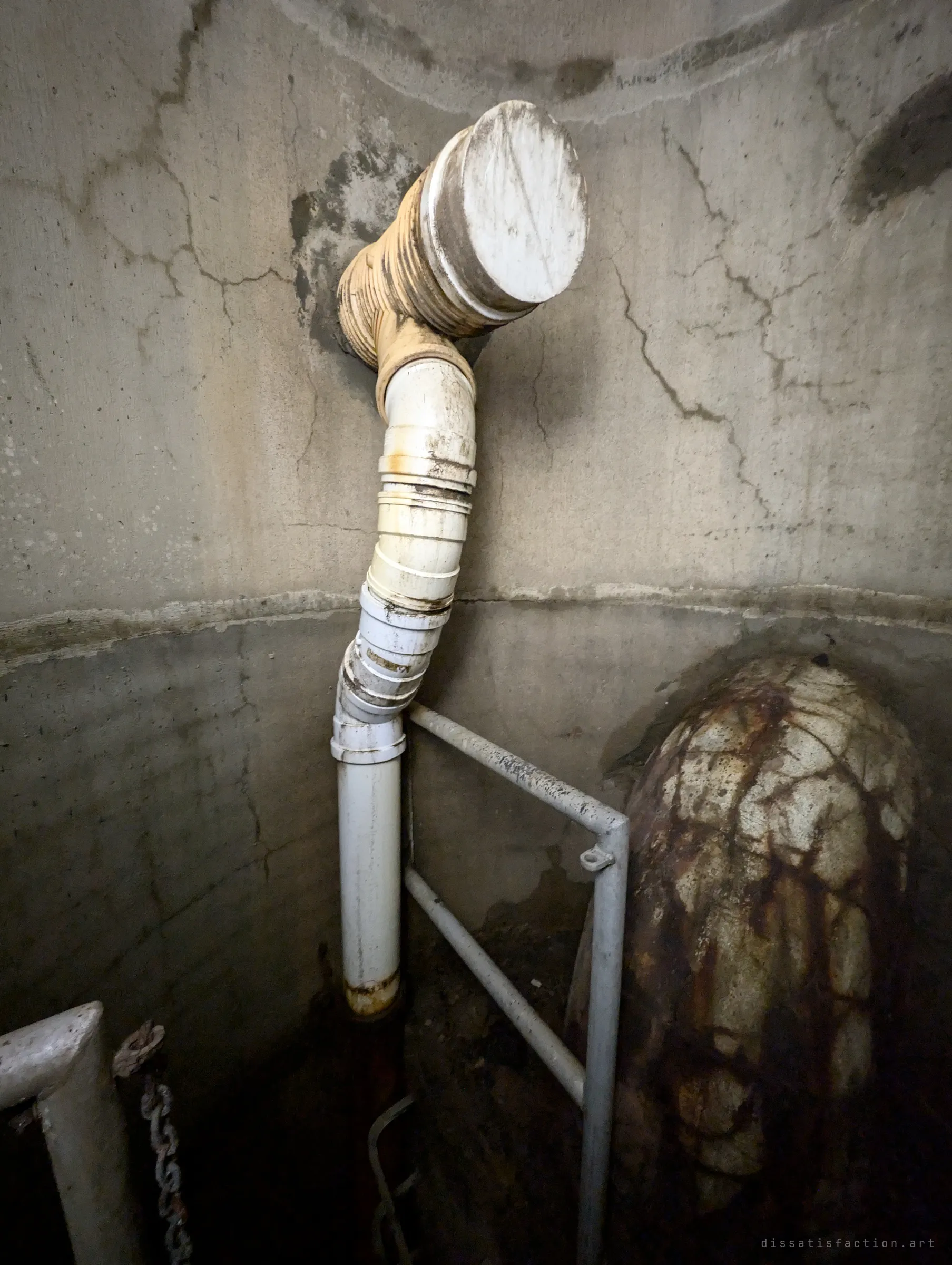 A patched-together plastic pipe snakes down a cracked concrete wall past a metal railing, beside a rust-streaked larger pipe entering the chamber. A close look at the improvised plumbing inside the drain.