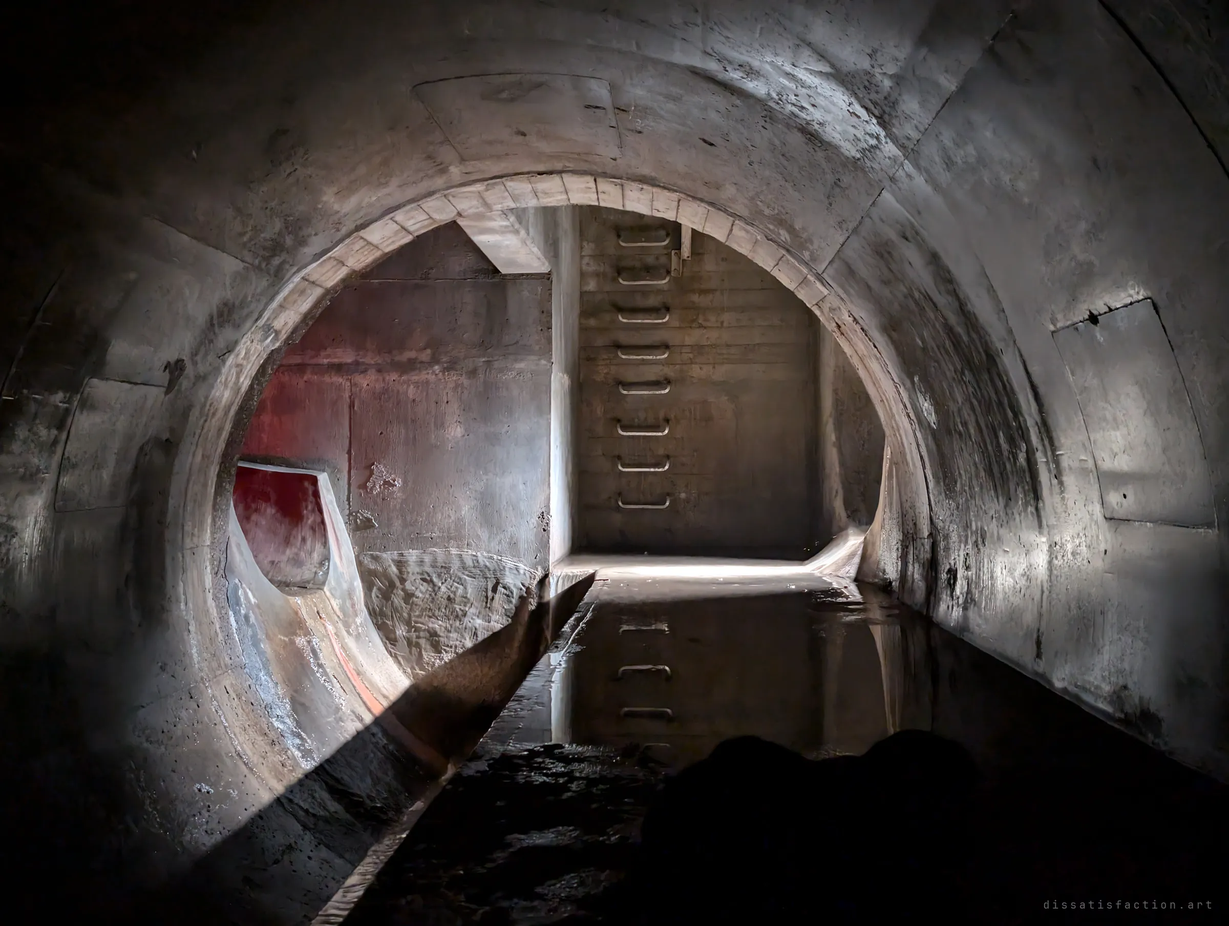 From inside a round pipe, the view opens onto a junction chamber with a ladder of step irons on the far wall, a red-lit side inlet and still water reflecting the rungs. The meeting point of several lines in the system.