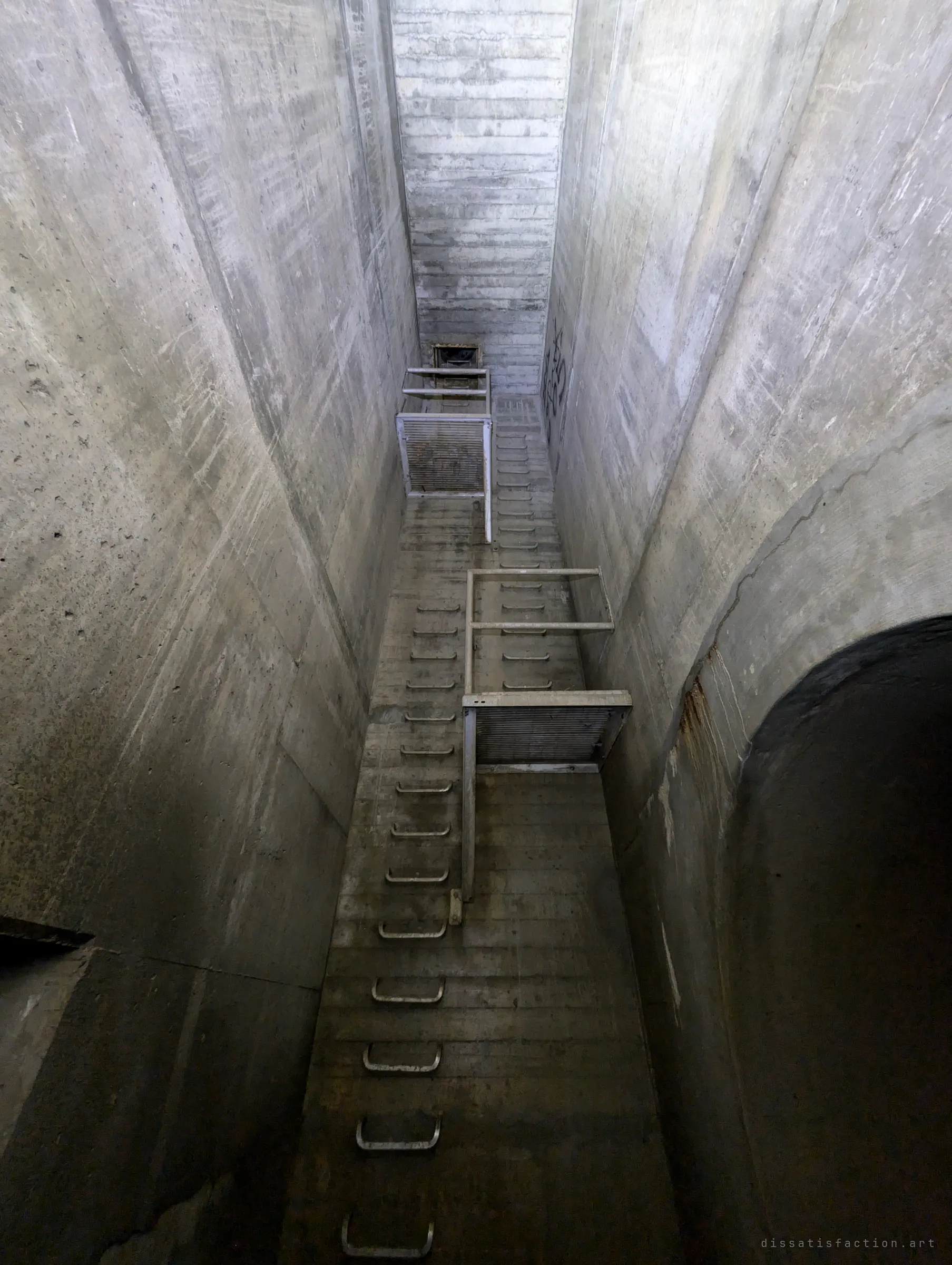 Looking straight up a tall, narrow concrete shaft lined with step irons and two caged landings toward a small grate at the top. Conveys the depth of the drain below the surface.