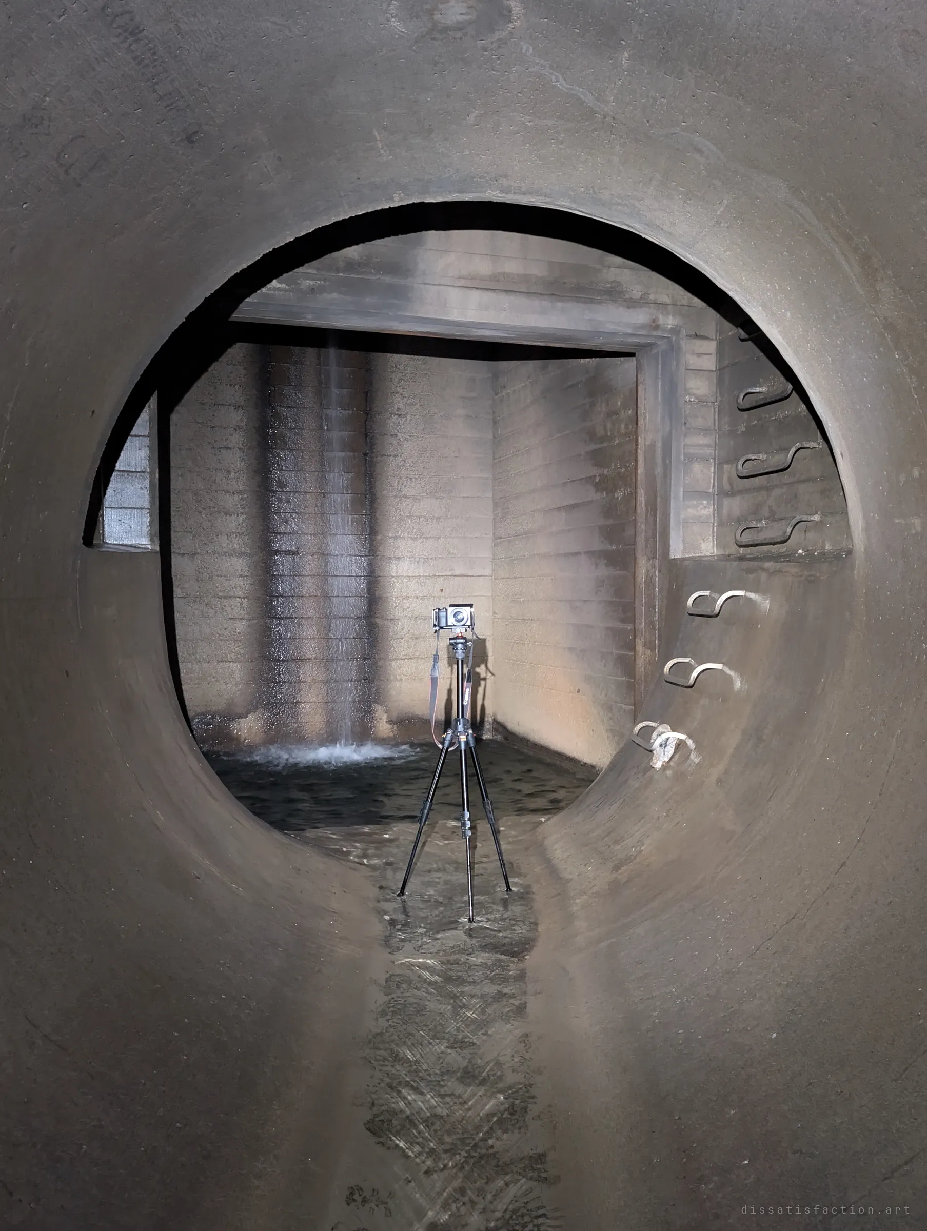 Framed by a circular pipe opening, a camera on a tripod stands in shallow water in front of a chamber where a thin stream pours down the back wall. Shows the photographer's setup at the plunge pool.