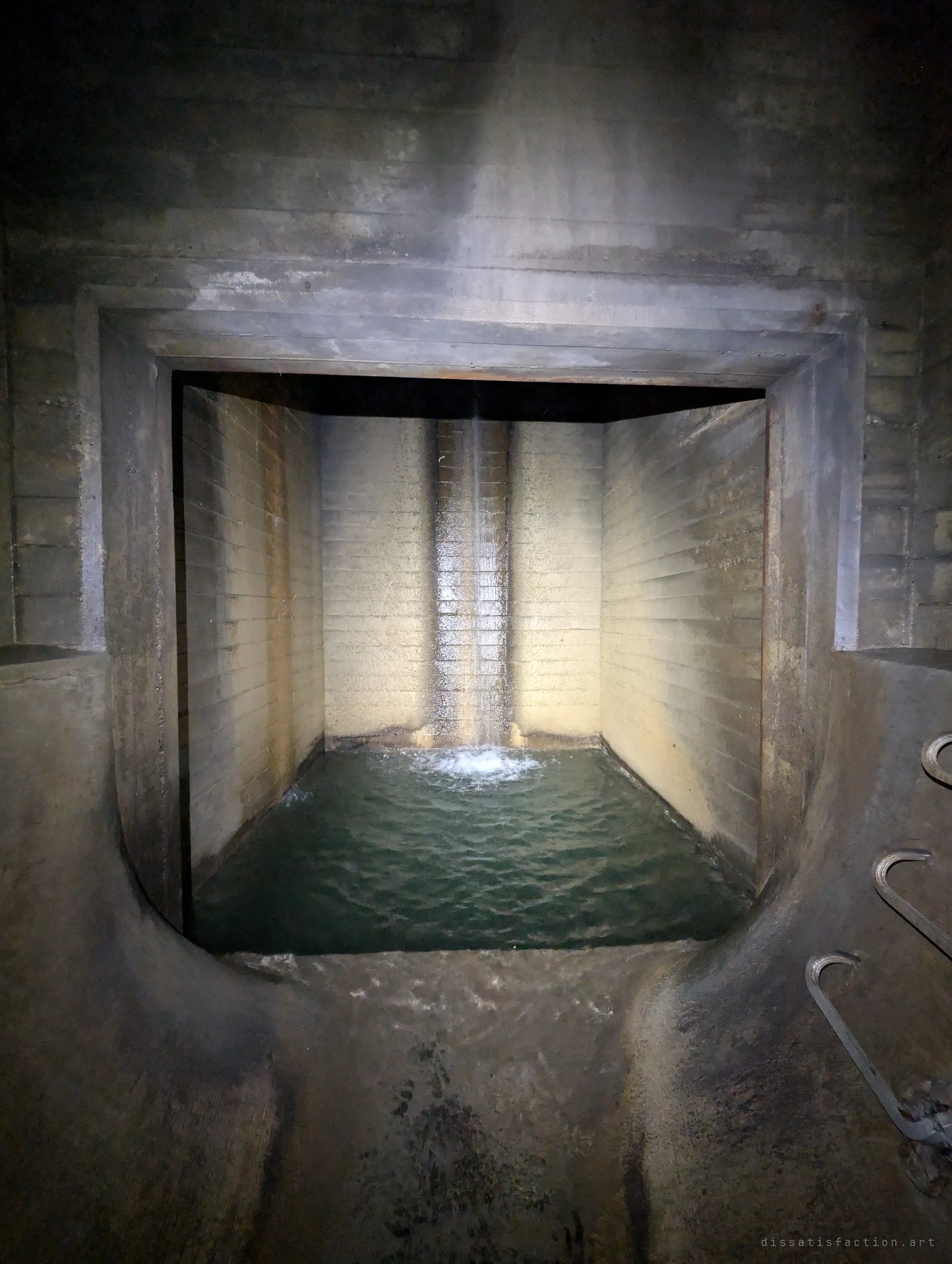 A square concrete plunge pool filled with green water, fed by a single stream falling from a slot in the back wall. It marks the furthest point reached in the drain.