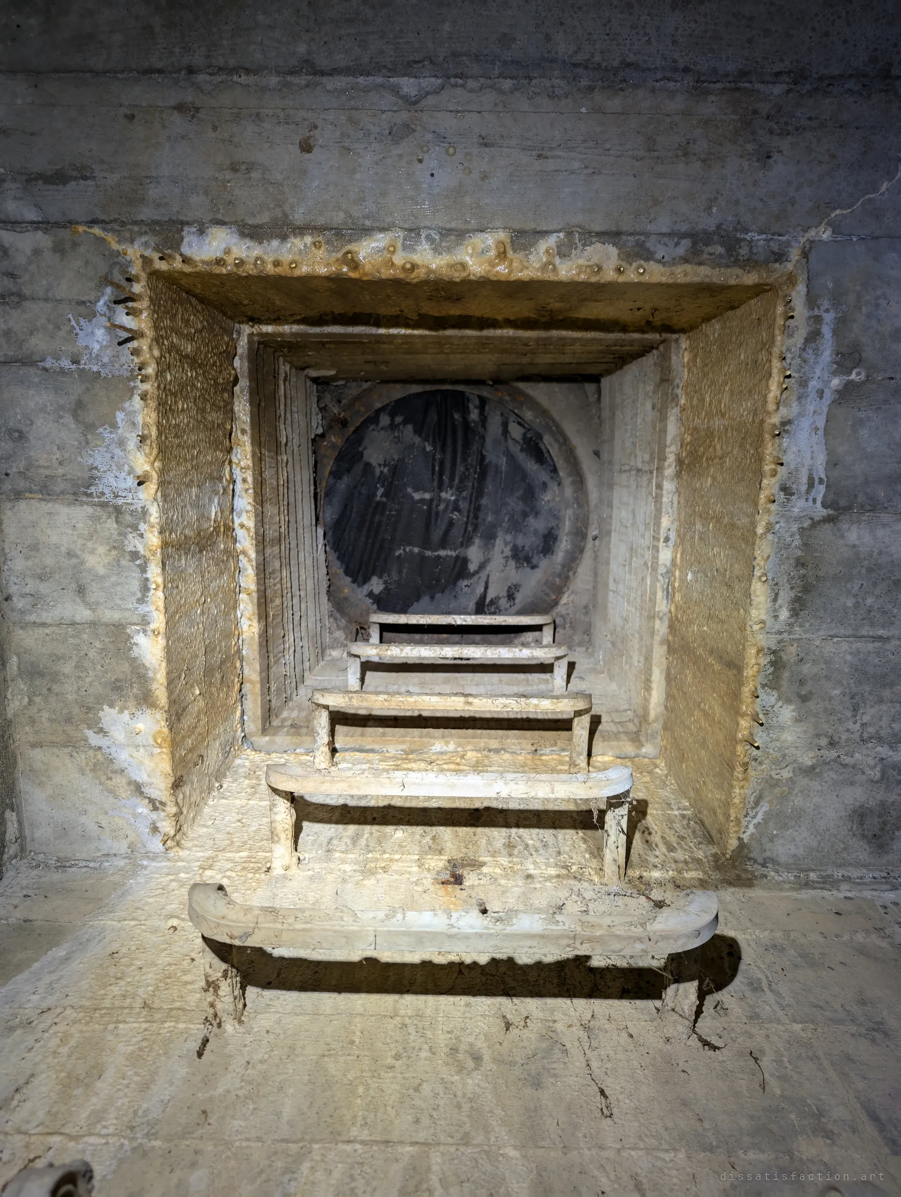 Looking up a square manhole shaft crusted with mineral deposits, with step rungs rising to a round lid. A stepped frame around the opening may catch debris falling from the street.