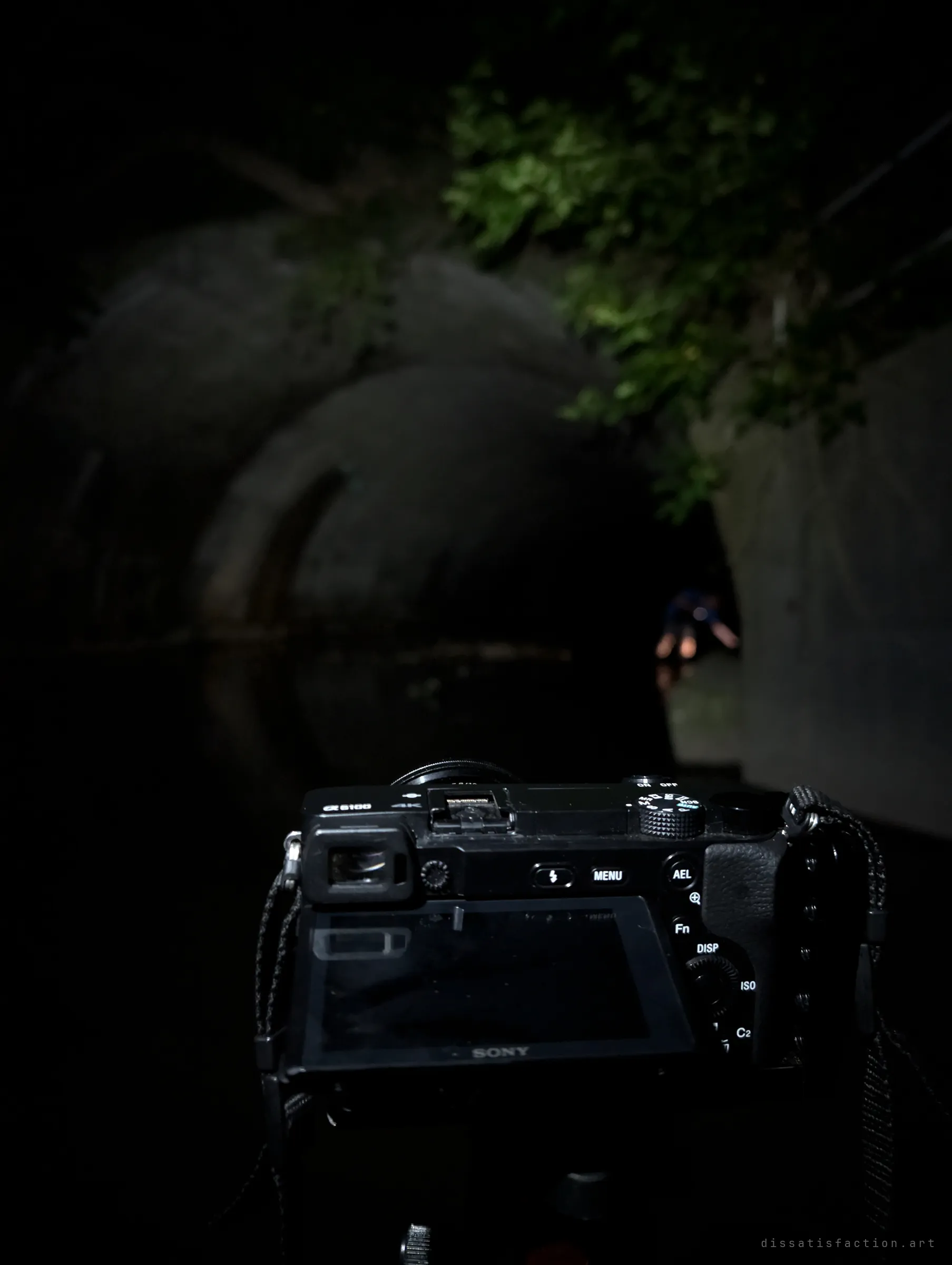 A mirrorless camera on a tripod faces into a dark concrete culvert mouth, vines hanging overhead and a distant explorer lit at the far end. A behind-the-scenes look at shooting in the dark.