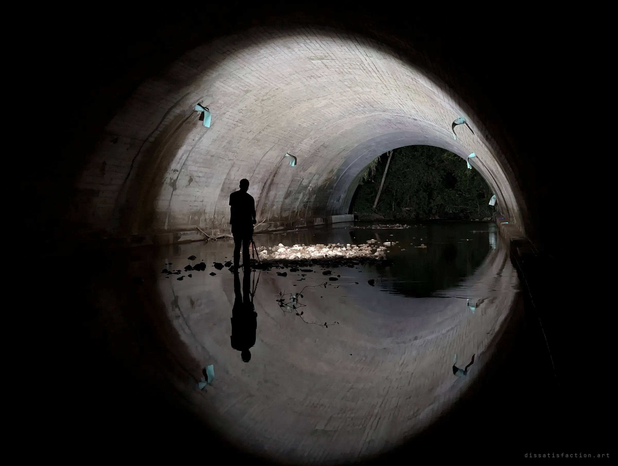 A silhouetted photographer with a tripod stands in shallow water inside a brick-lined arched culvert, the flashlight beam turning the tunnel and its reflection into a near-perfect ring, with trees at the far opening.