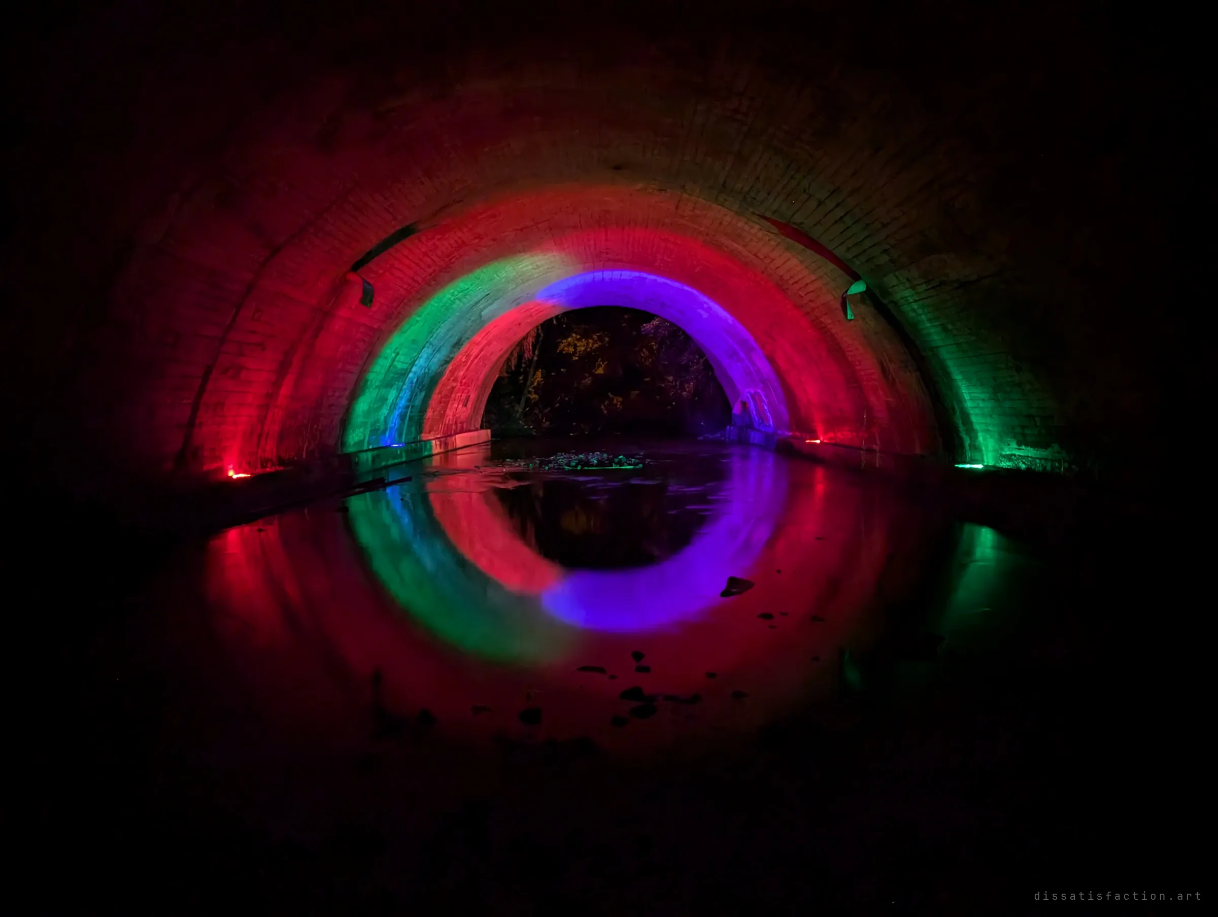 Red, green and violet lights paint concentric arches along a brick culvert, doubled into a glowing ring by the still water below.