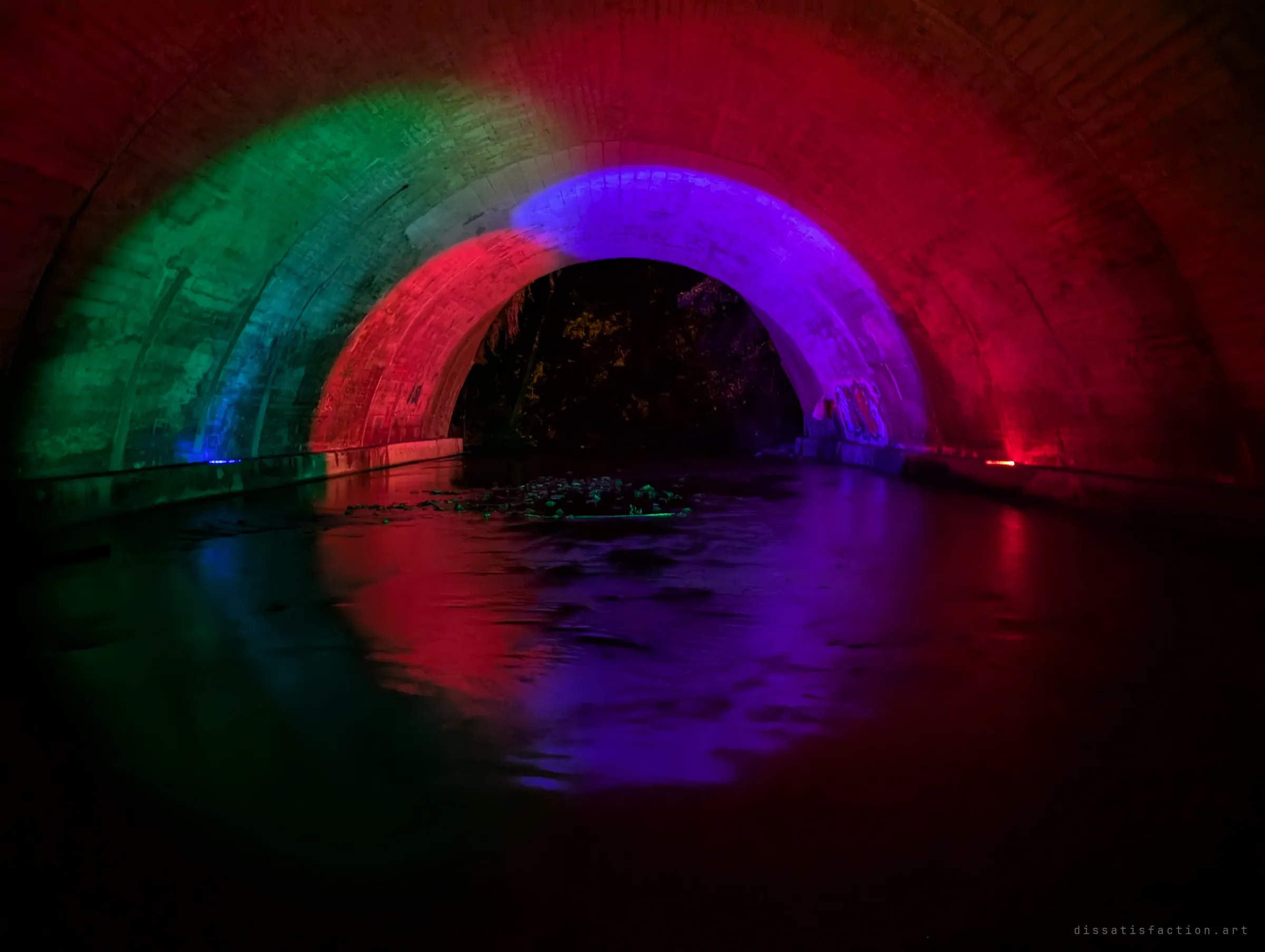A closer view of the light-painted culvert: green, red and violet bands sweep the brick arches, with small graffiti tags near the far opening and coloured reflections rippling on the water.