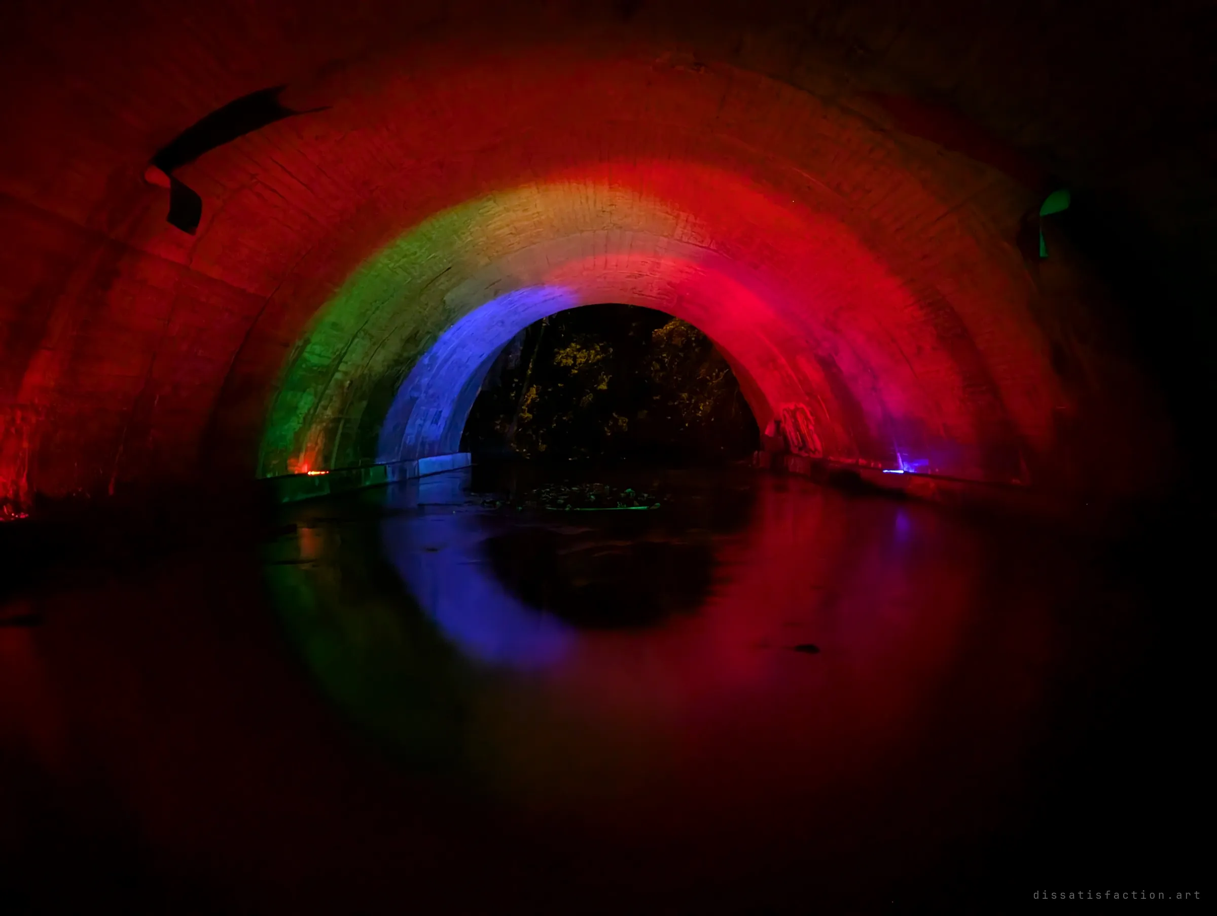 A third lighting pass through the culvert, a rainbow of red, green and blue arching over dark water toward the leafy exit.
