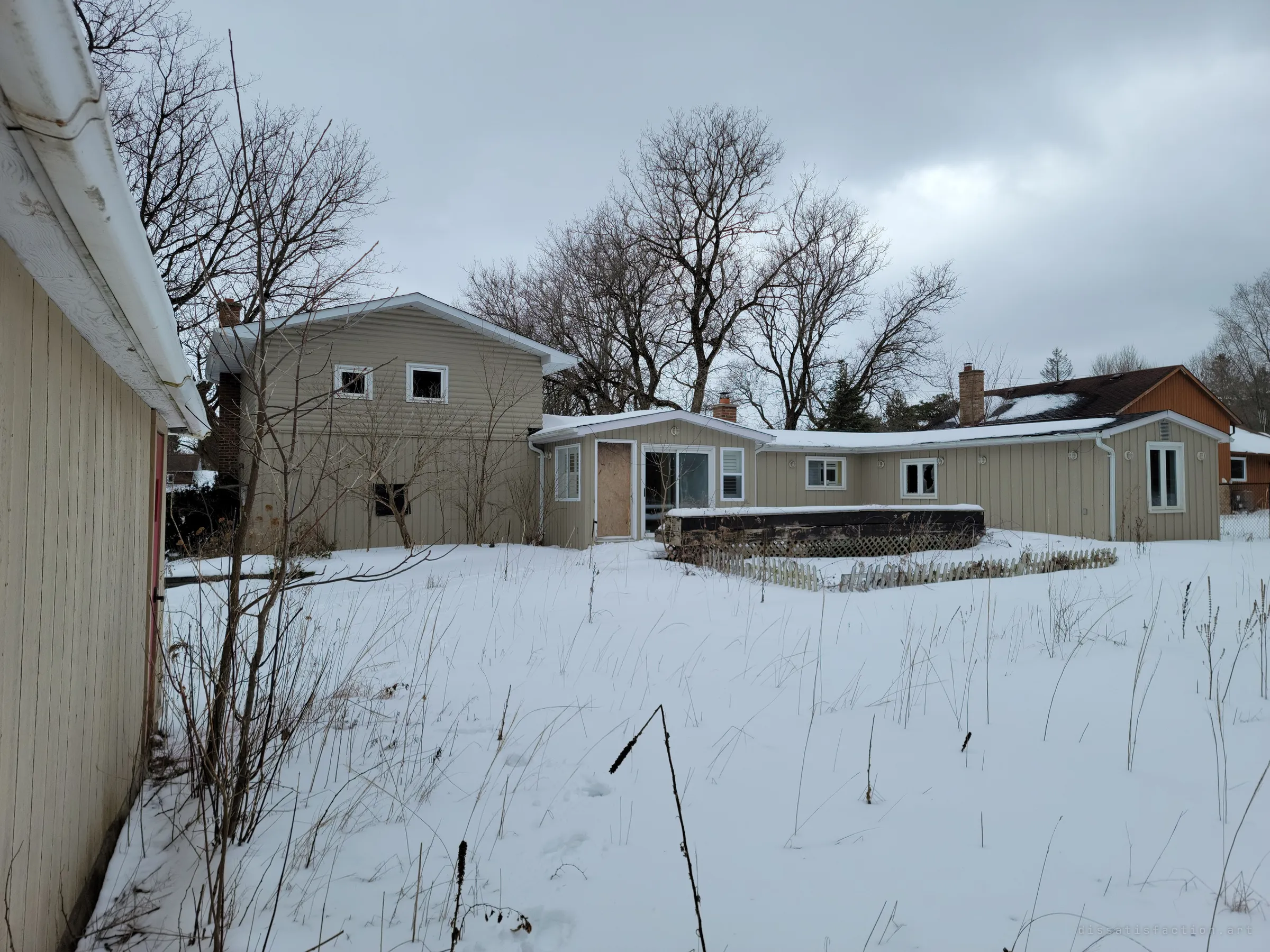 The back of the house under grey skies, with a boarded door, a snow-buried deck and a neighbouring home beyond. Tall dry grass pokes through the snow of the overgrown yard.