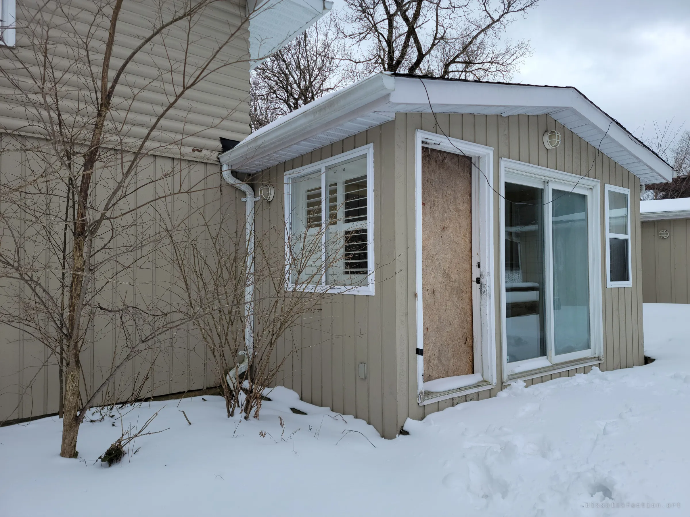 A small sunroom addition with its door boarded over in plywood and a sliding glass door beside it. A loose wire hangs from the roof edge above the snow.