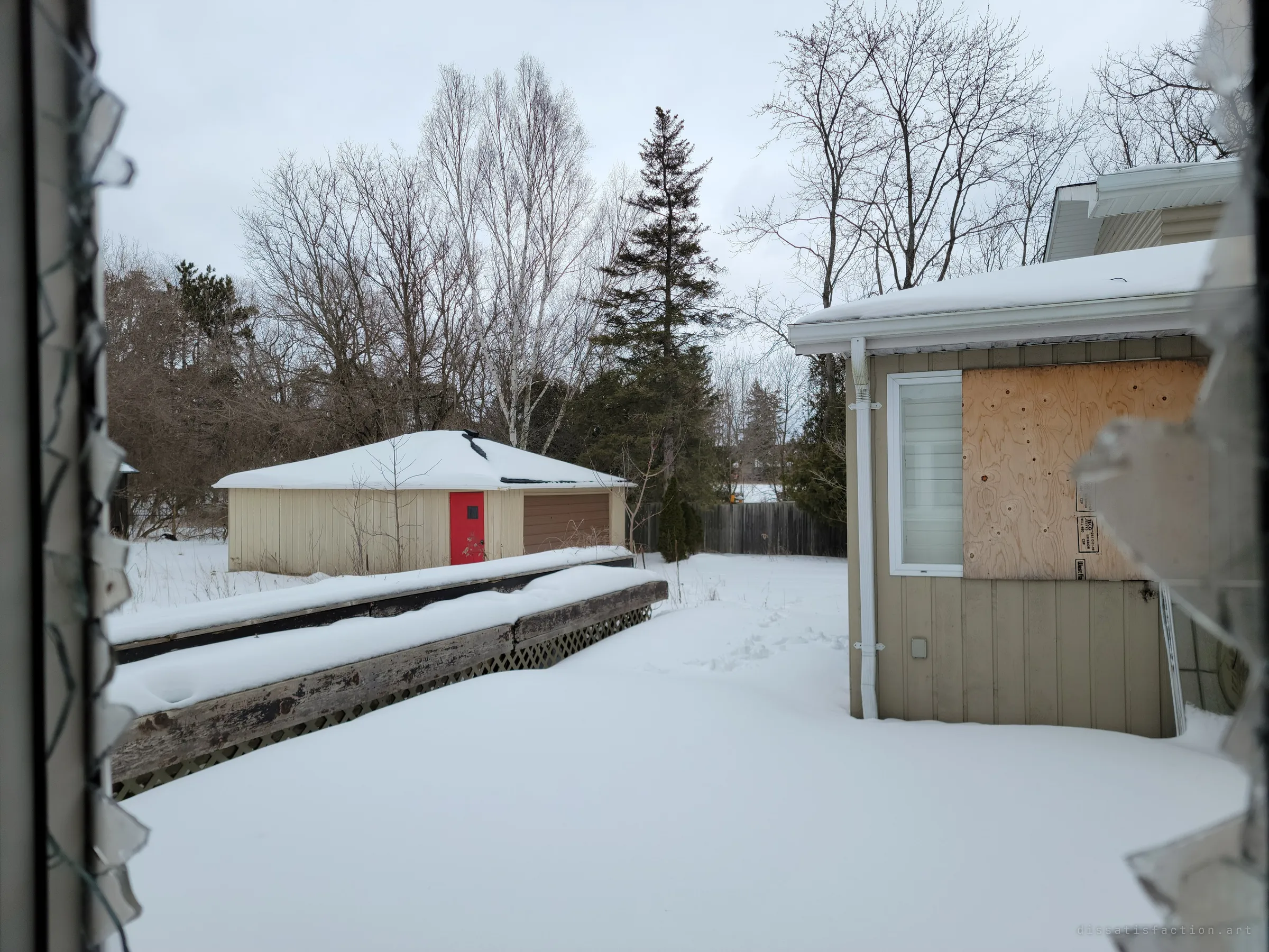 Looking out through a shattered window at a detached garage with a bright red door and a snowed-in deck. Jagged glass shards frame the view on both sides.