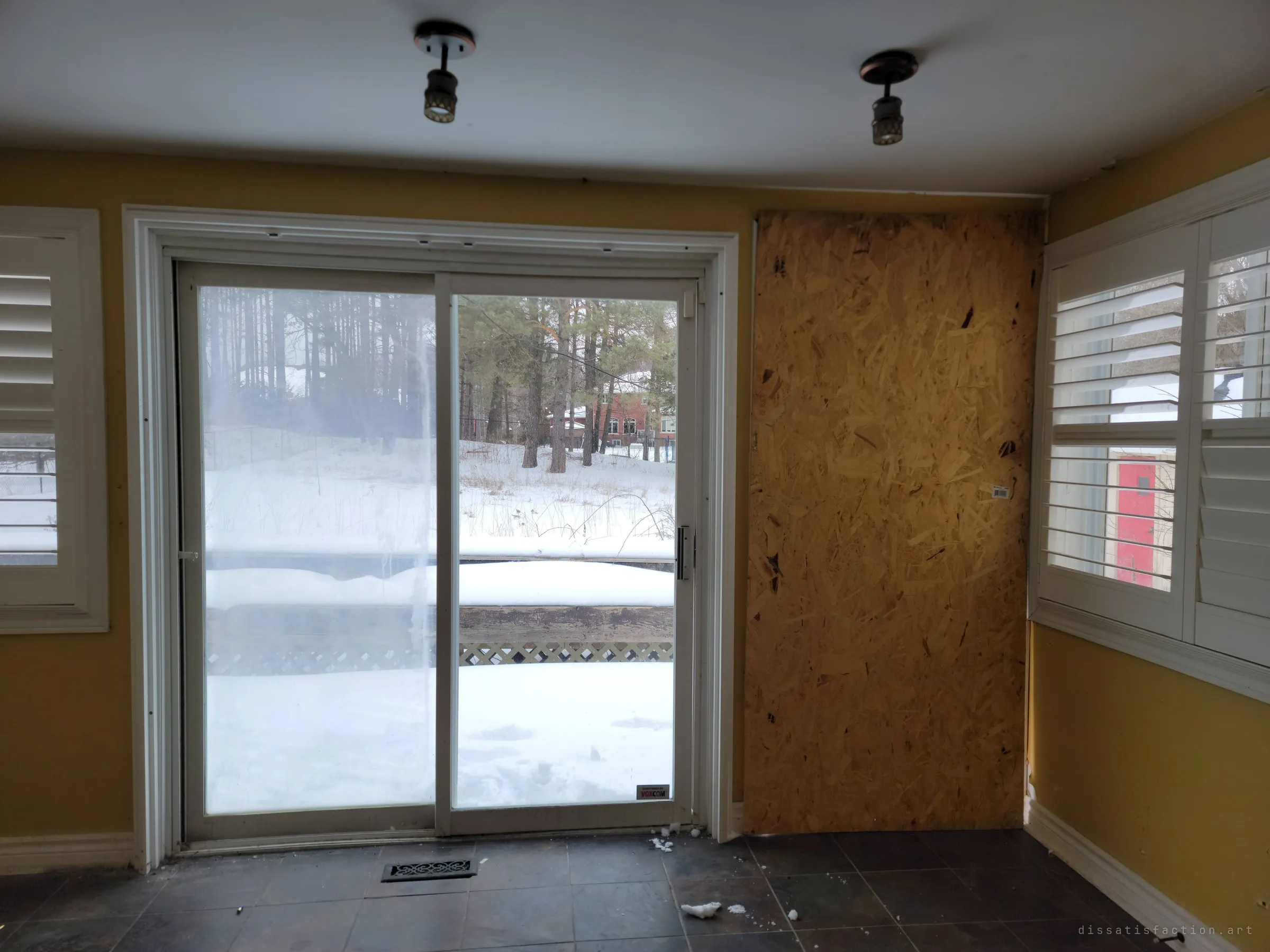 Inside the sunroom, a sliding patio door looks out over a snowy yard into pine trees, next to a door sealed with plywood. Shuttered windows and yellow walls still look cared for.