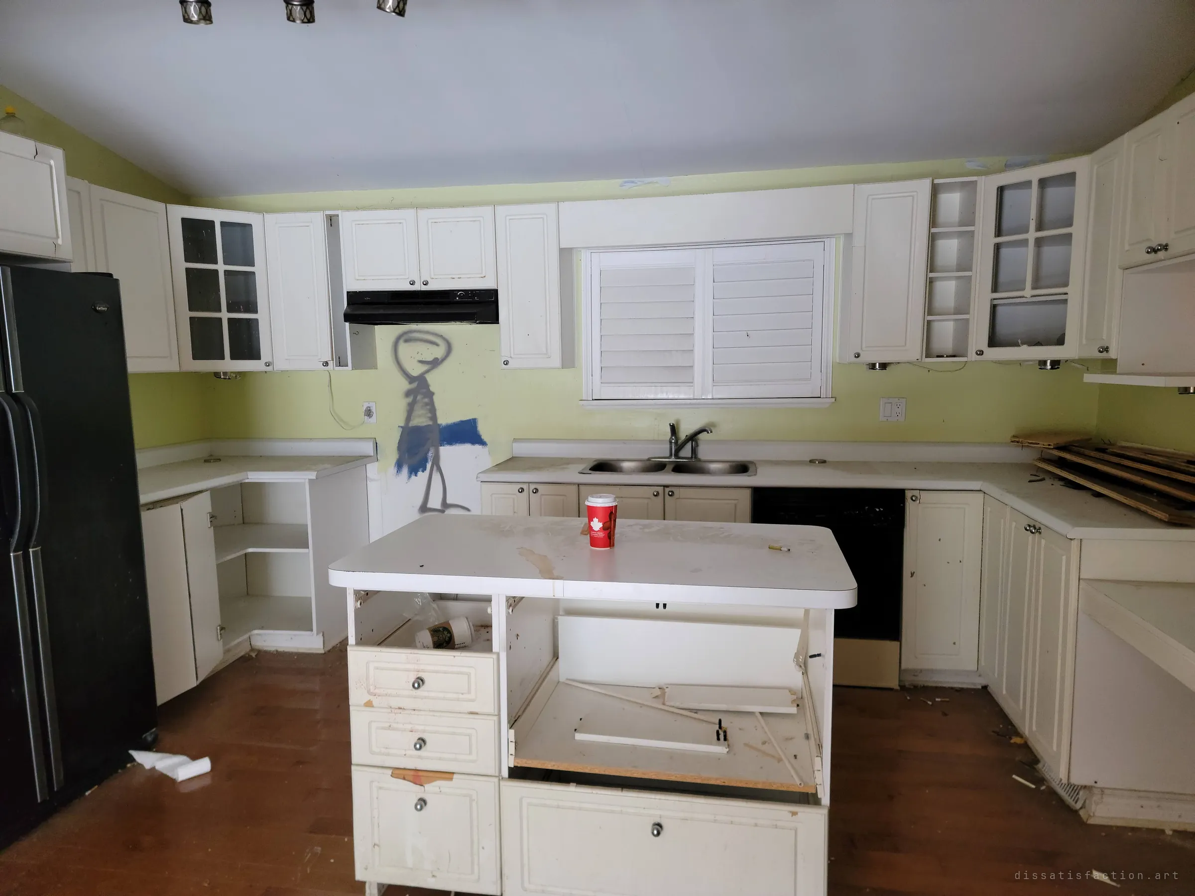 A gutted kitchen with doors and drawers torn from the island and a spray-painted stick figure on the wall. A coffee cup left on the counter hints at recent visitors.