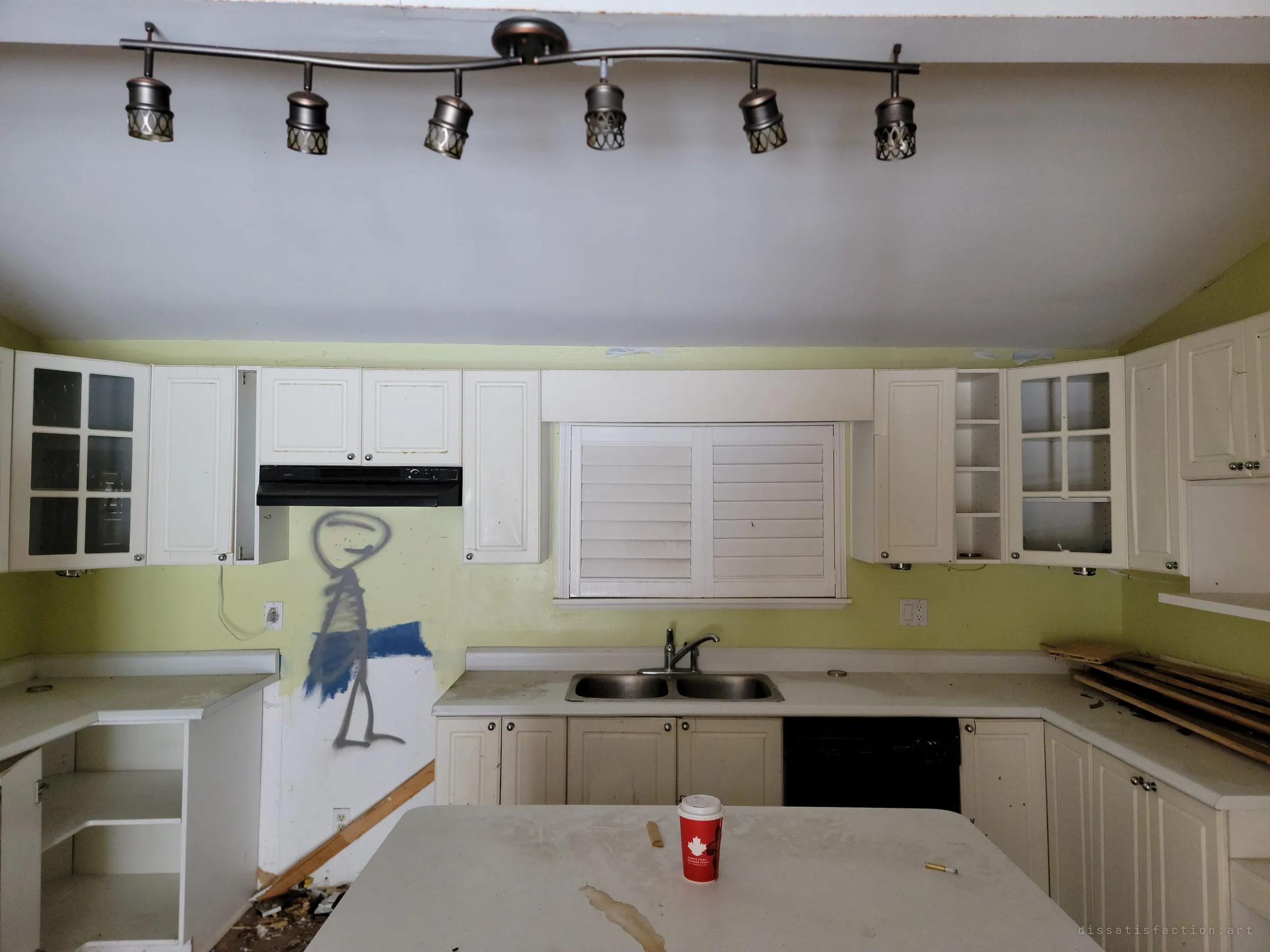 A straight-on view of the same kitchen under a track light, with the stick-figure graffiti beside the range hood. Wood debris is piled on the counters and a coffee cup sits on the island.
