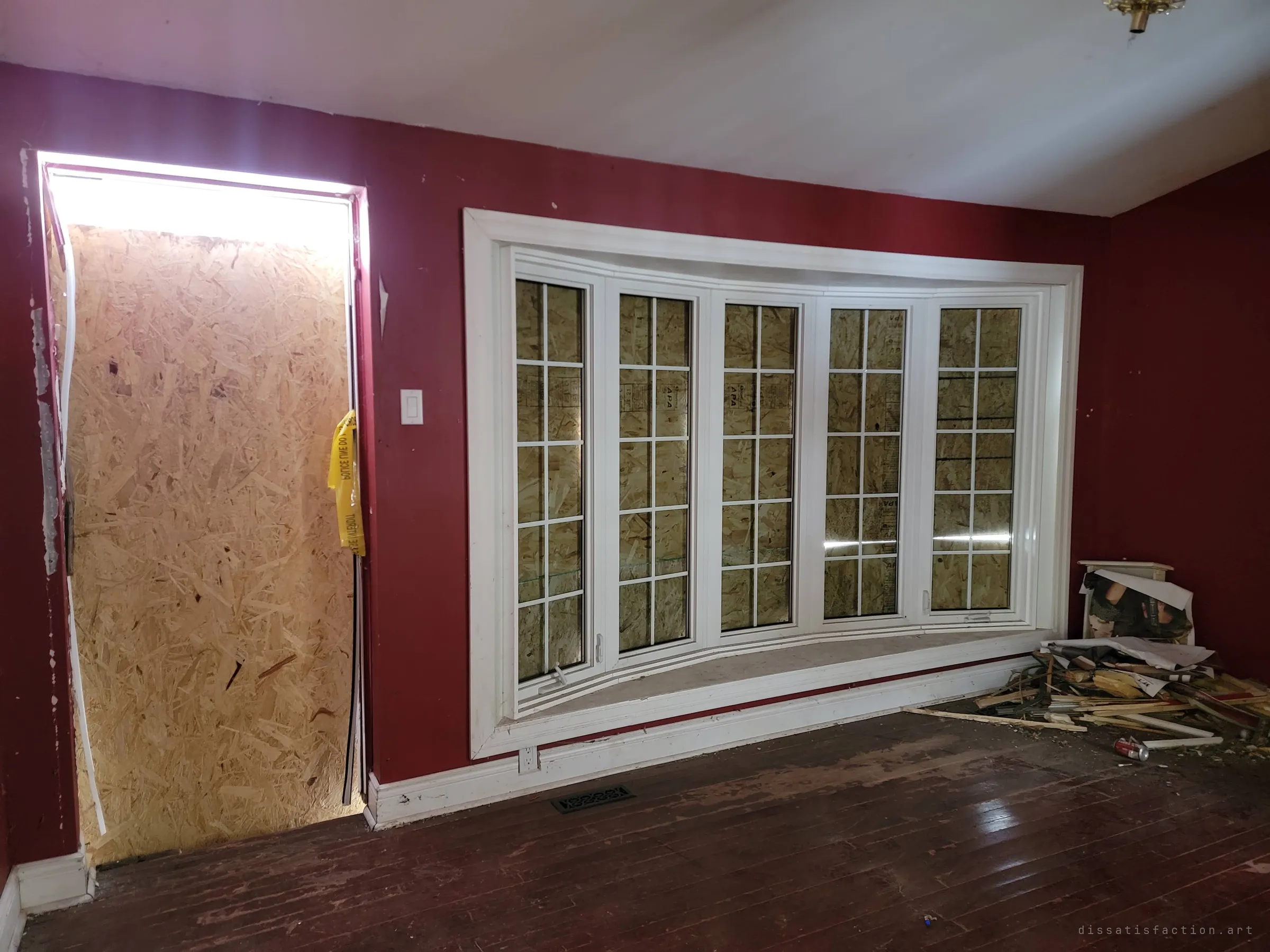A deep red room with a bay window and front door boarded from outside, daylight leaking around the plywood. Torn caution tape hangs by the door and debris is heaped in the corner.
