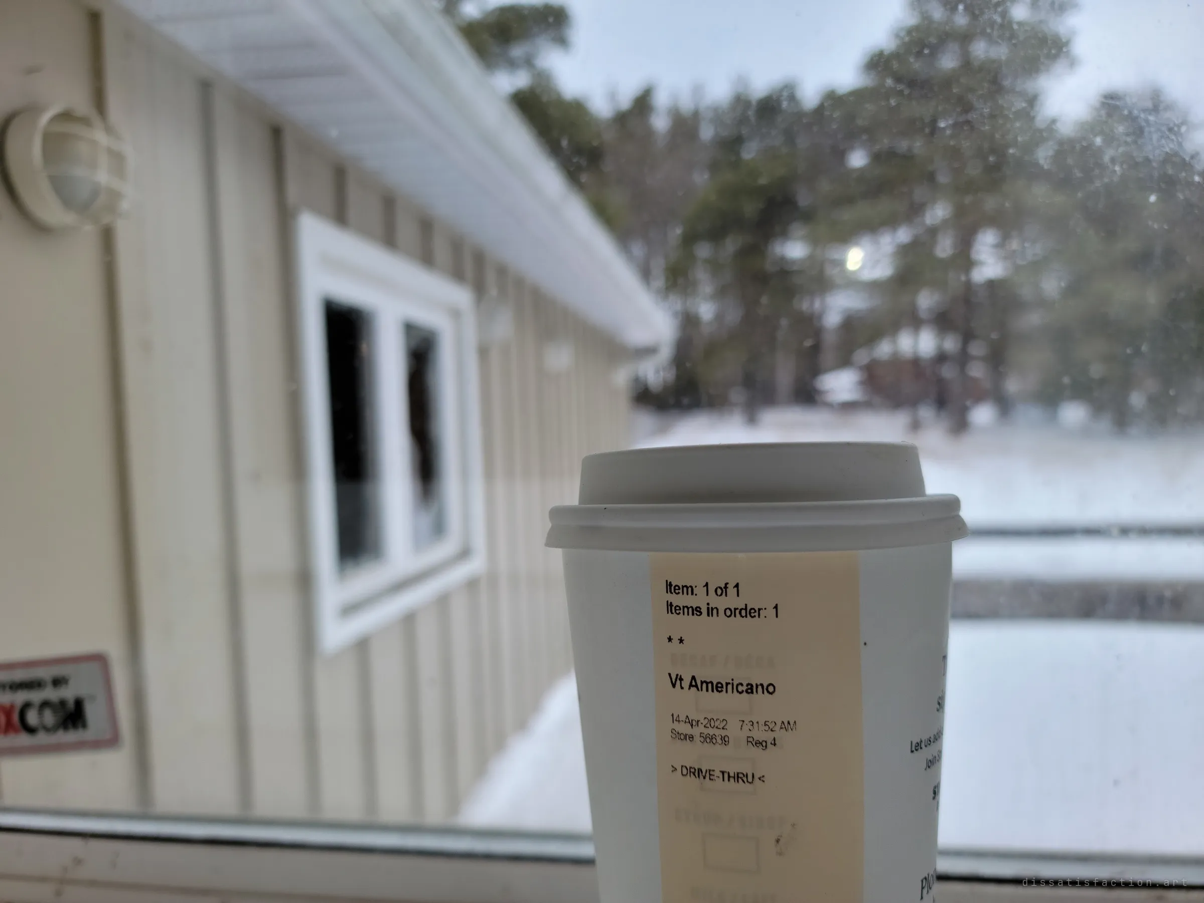 A takeaway coffee cup held up at a window, with the house's siding and snowy pines blurred behind it. Its order sticker is a small trace of someone passing through.