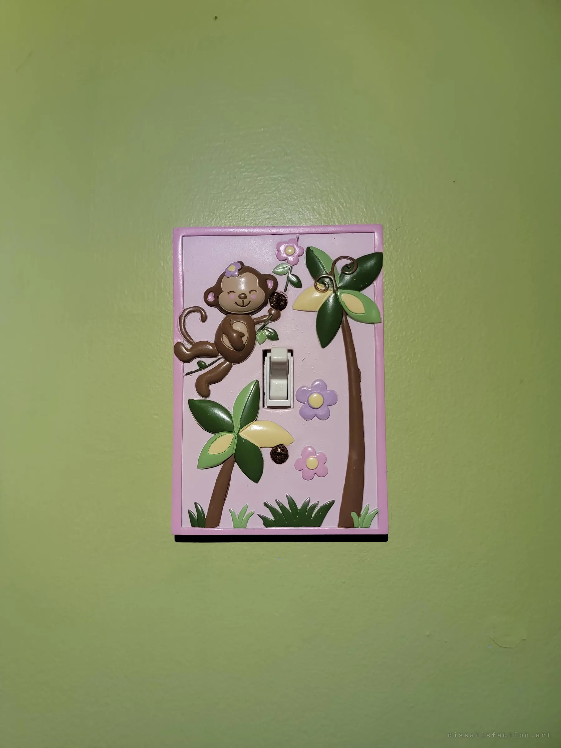 A pink light-switch plate moulded with a smiling monkey, palm trees and flowers on a green wall. A small leftover of the child's room that survived the decay.