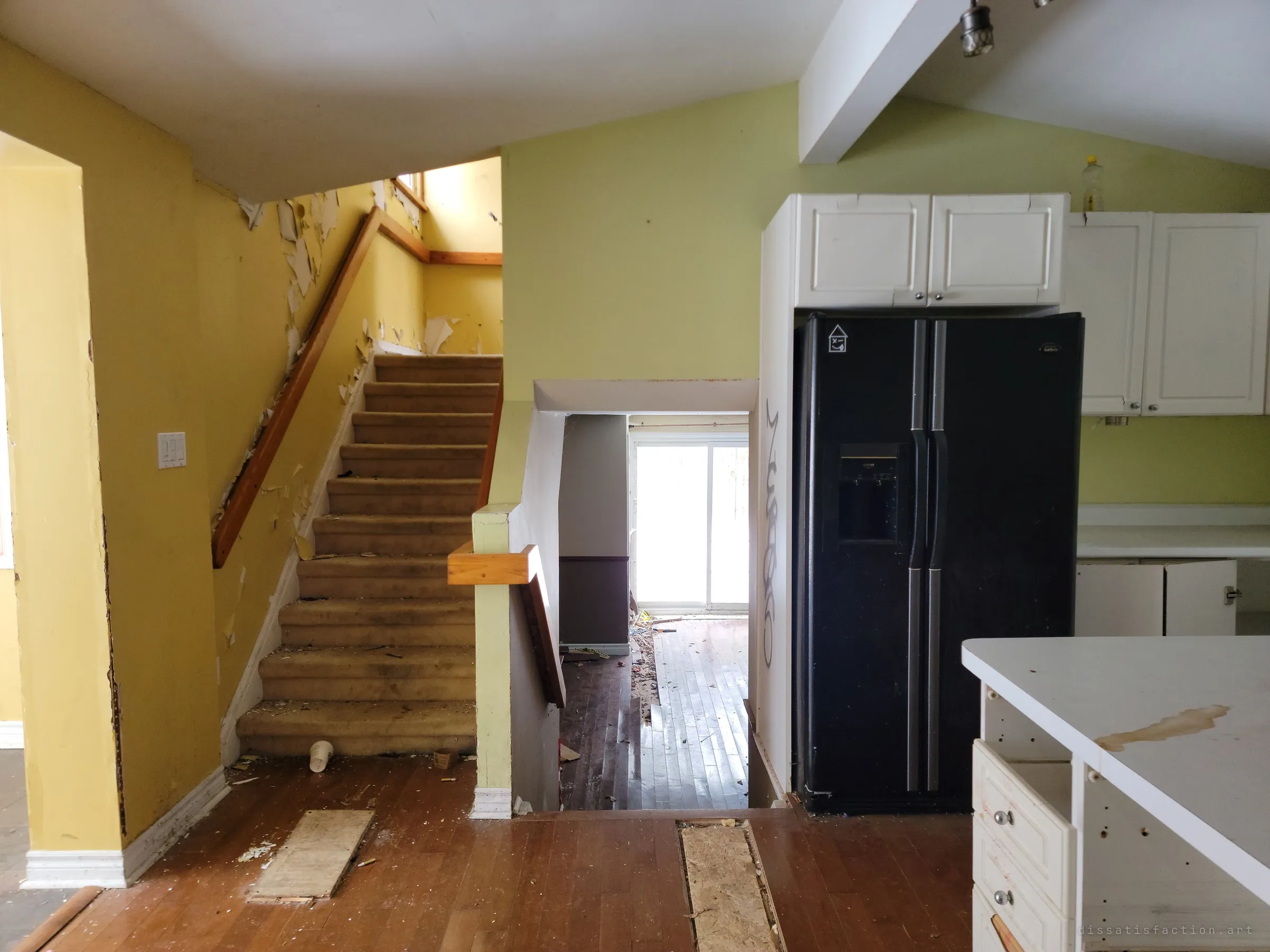 An open-plan kitchen with a tagged black fridge, a stained island and carpeted stairs rising past peeling yellow walls, with a split-level drop to a bright back room. The layout of a lived-in family home, now gutted.